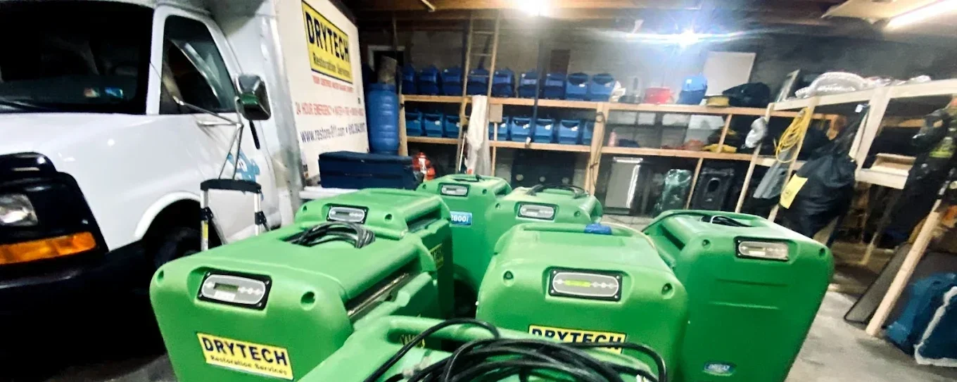 Multiple green DRYTECH dehumidifiers in a storage space with shelves holding blue and black containers, tools, and other equipment.
