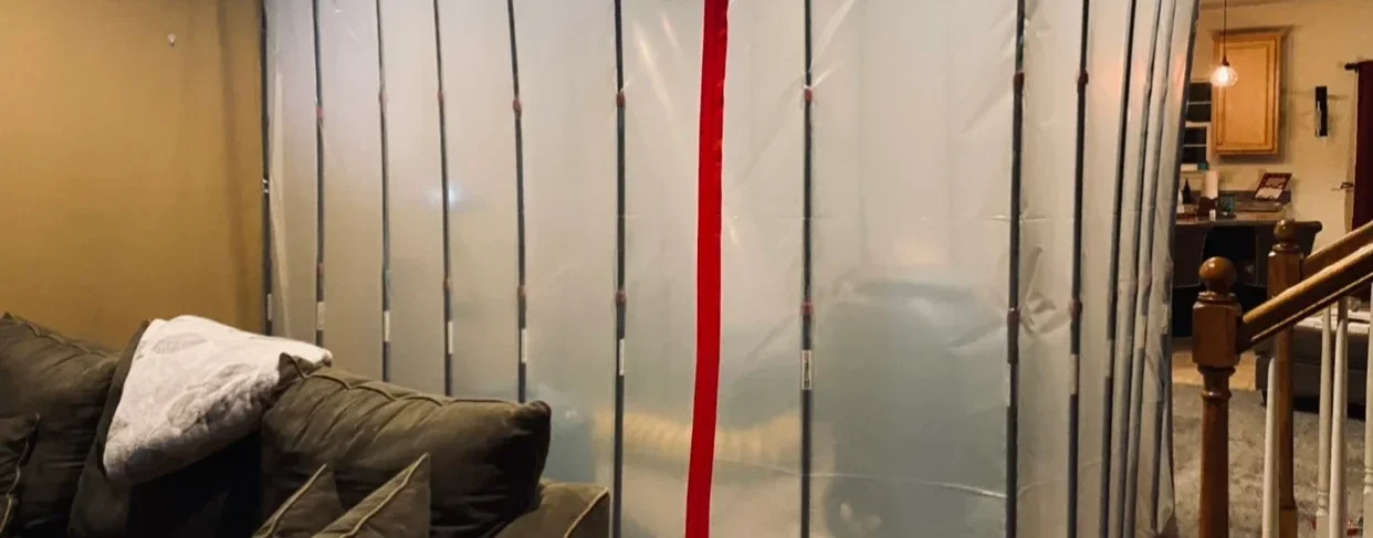 A plastic sheet taped across a doorway, with a red strip of tape in the middle, partially obscuring the view of a living room.