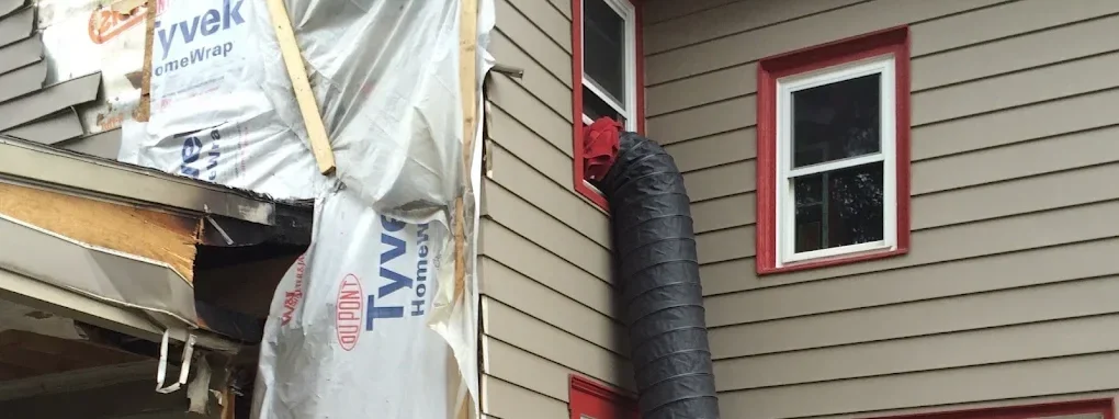 A house under construction or renovation with new vinyl siding being installed. A worker's arm is visible inside a red-framed window, and a large black flexible duct is attached to the house exterior. Building materials and insulation are visible on 