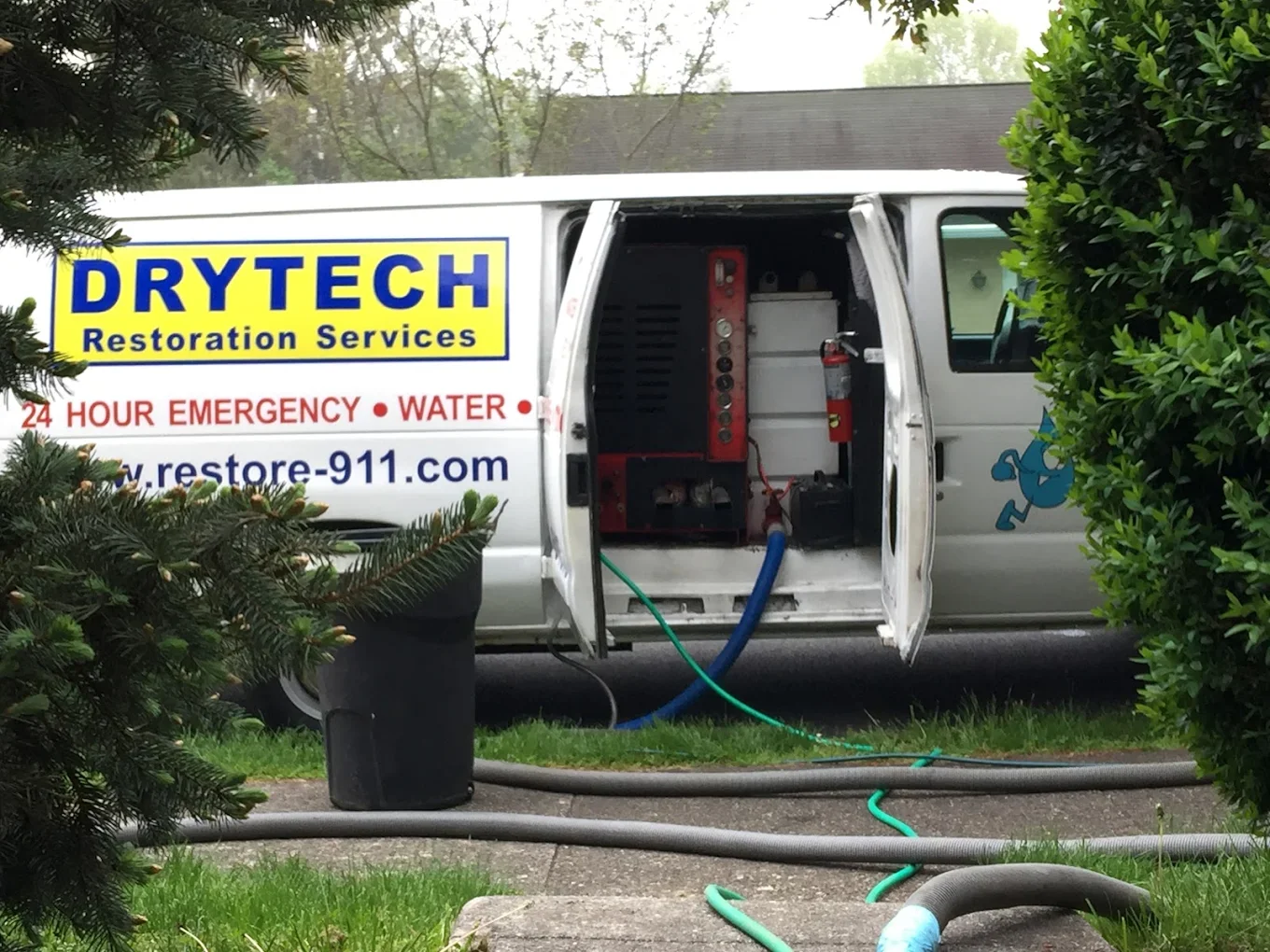 A white service van labeled 'DRYTECH Restoration Services' with open side doors revealing equipment inside; hoses and tools are on the ground nearby, and it is parked on a driveway surrounded by greenery and trees.