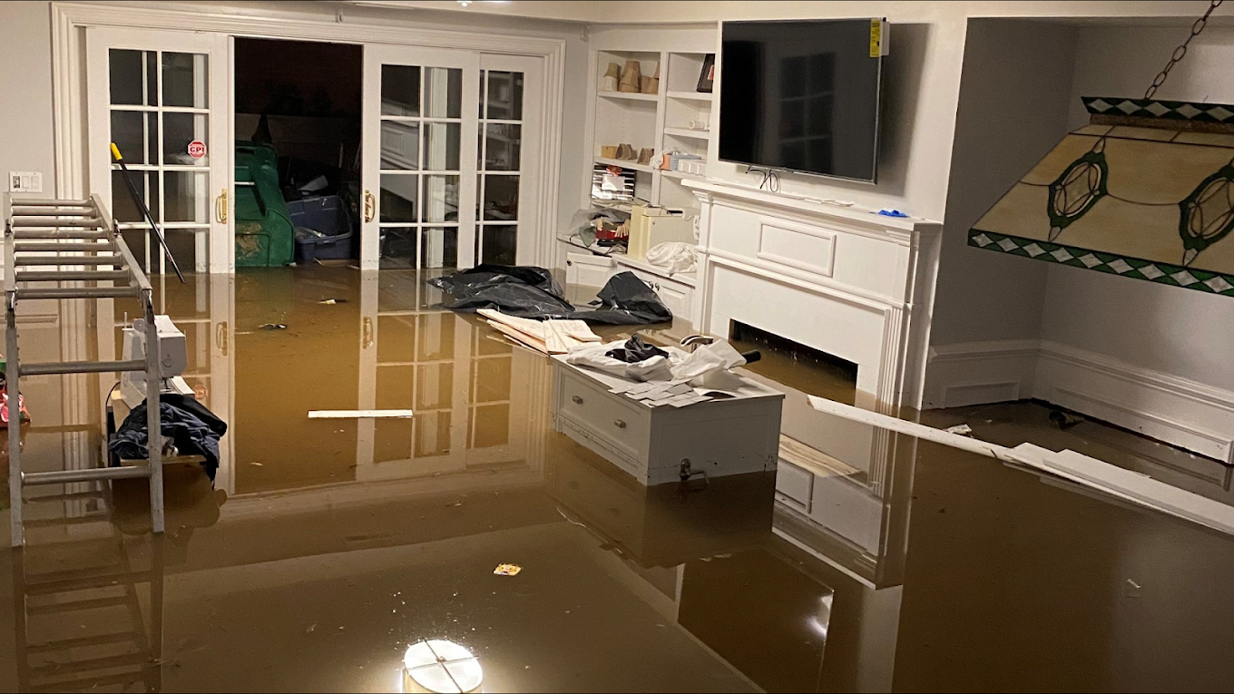 Living room heavily flooded with muddy water, partially submerged furniture and cluttered items, visible through glass sliding doors, indicating recent flood damage.