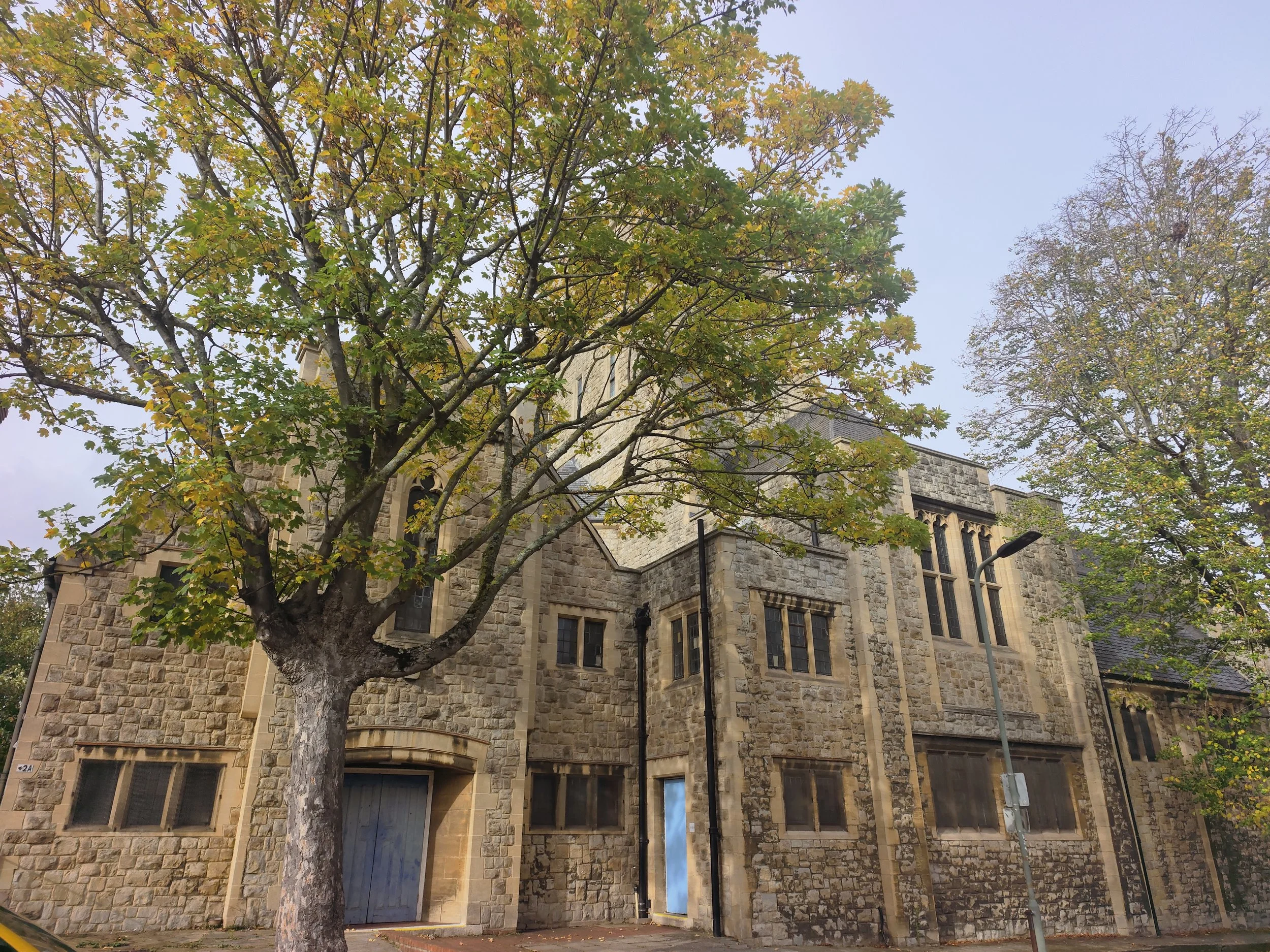 Penge Congregational Church.. Stone church with many windows, surrounded by trees with green and yellow leaves, on a cloudy day.