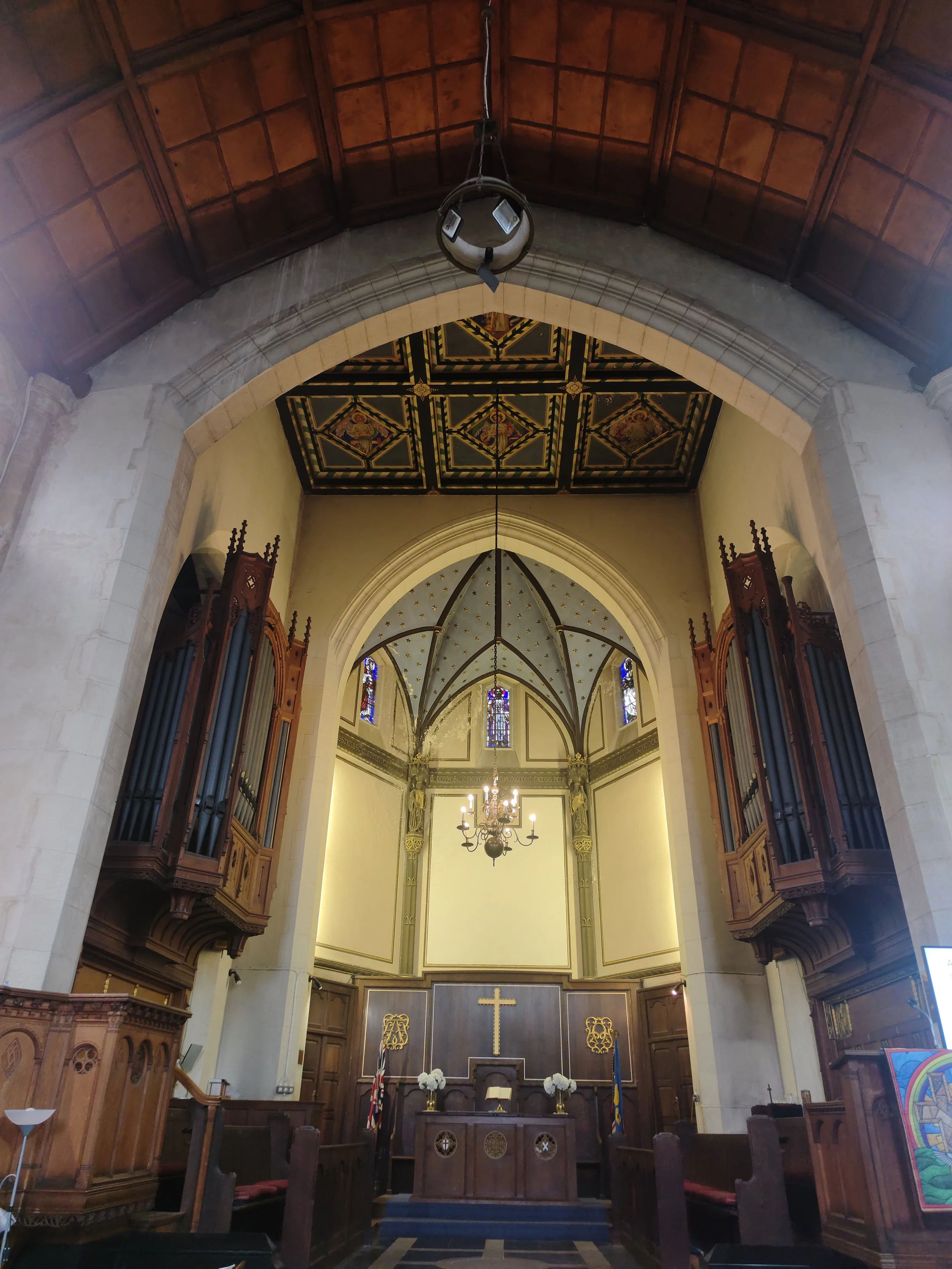Penge Congregational Church. Interior of a church altar with a cross, altar table, flower arrangements, stained glass windows, a chandelier, and wooden organ pipes on each side.
