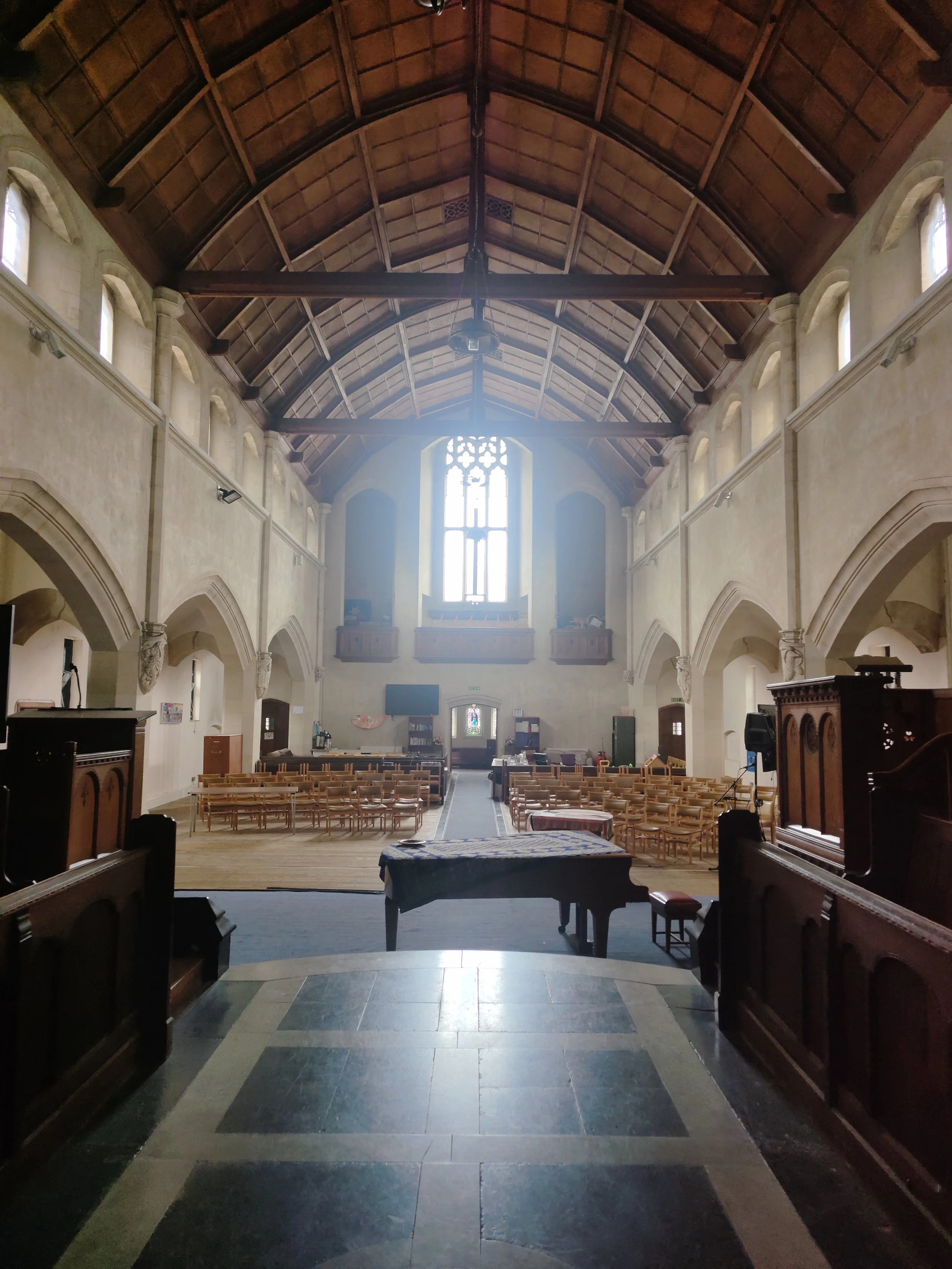 Penge Congregational Church. Interior of a church with a high wooden vaulted ceiling, rows of wooden chairs, a grand piano, and a large stained glass window at the front.