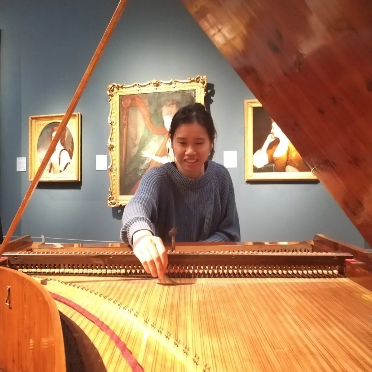A woman playing a grand piano in an art gallery with framed paintings on the wall behind her.