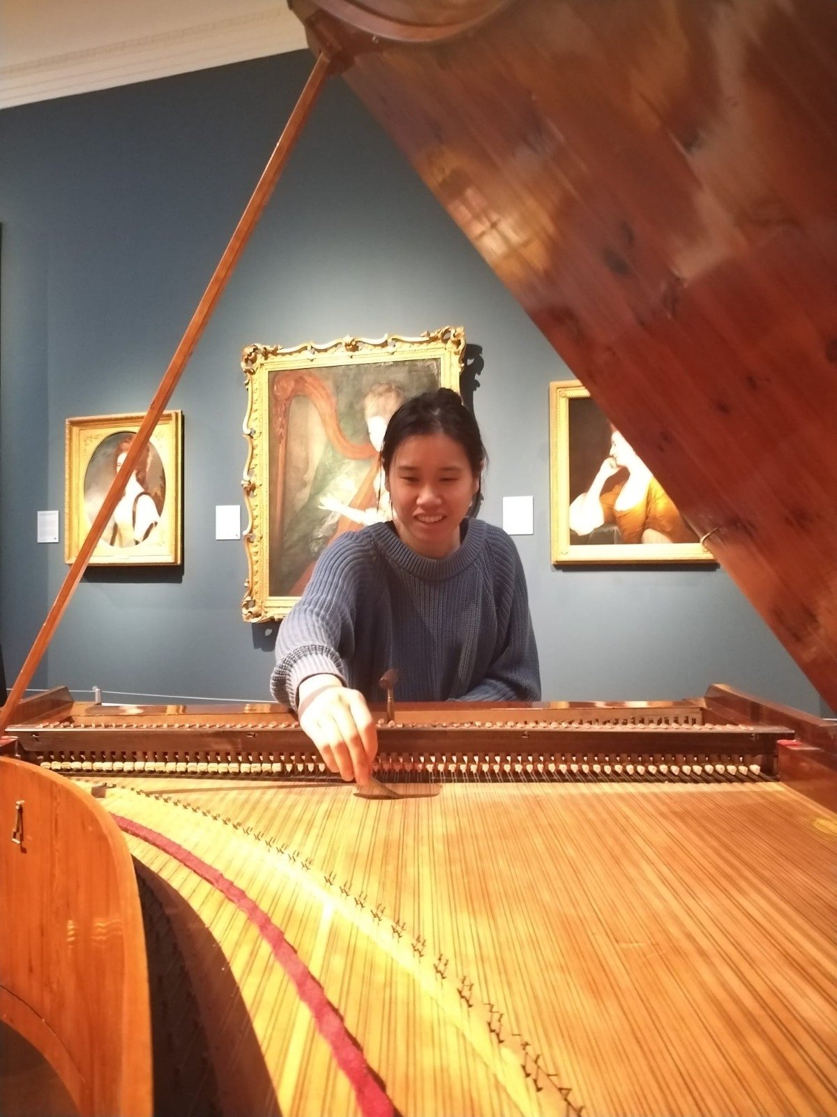 A woman in a grey sweater playing an open, wooden grand piano in an art gallery, with paintings hanging on a dark teal wall in the background.