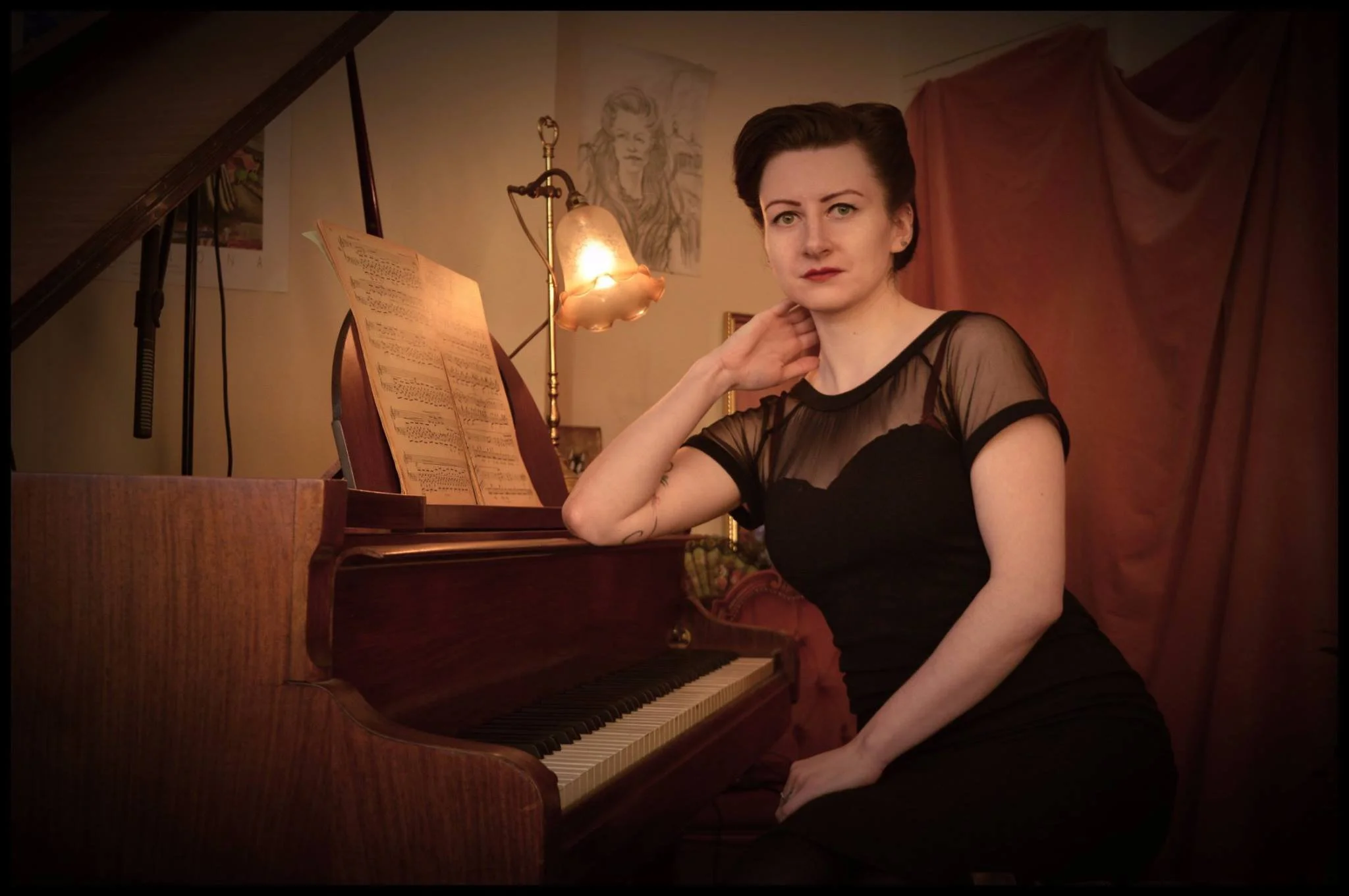 A woman with short dark hair wearing a black dress with a sheer top, sitting at a vintage upright piano with sheet music, in a warmly lit room decorated with framed pictures and a lamp.