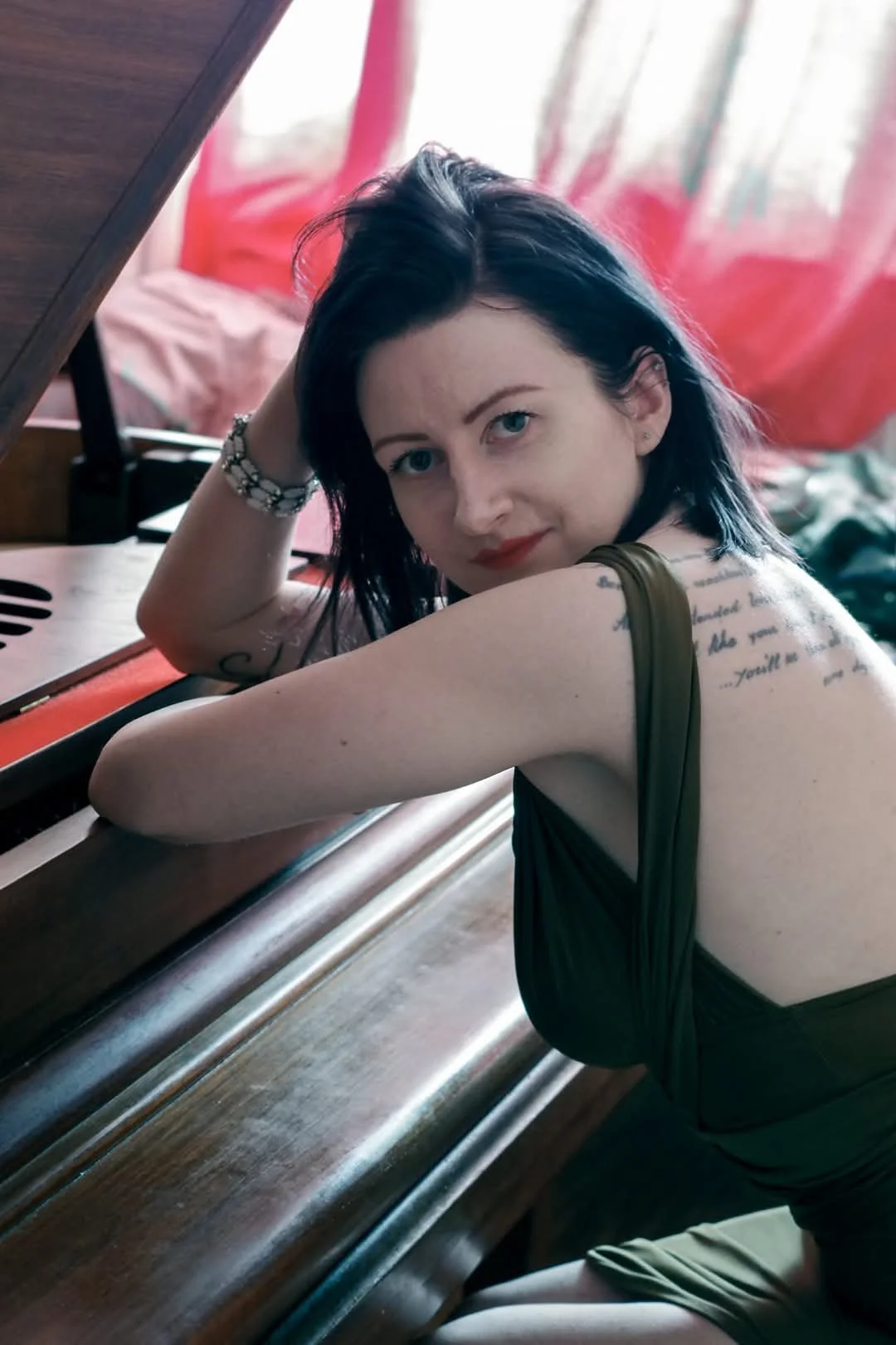 A young woman with dark hair and tattoos on her arm sitting at a piano, resting her head on her arm, looking at the camera, in a softly lit room.