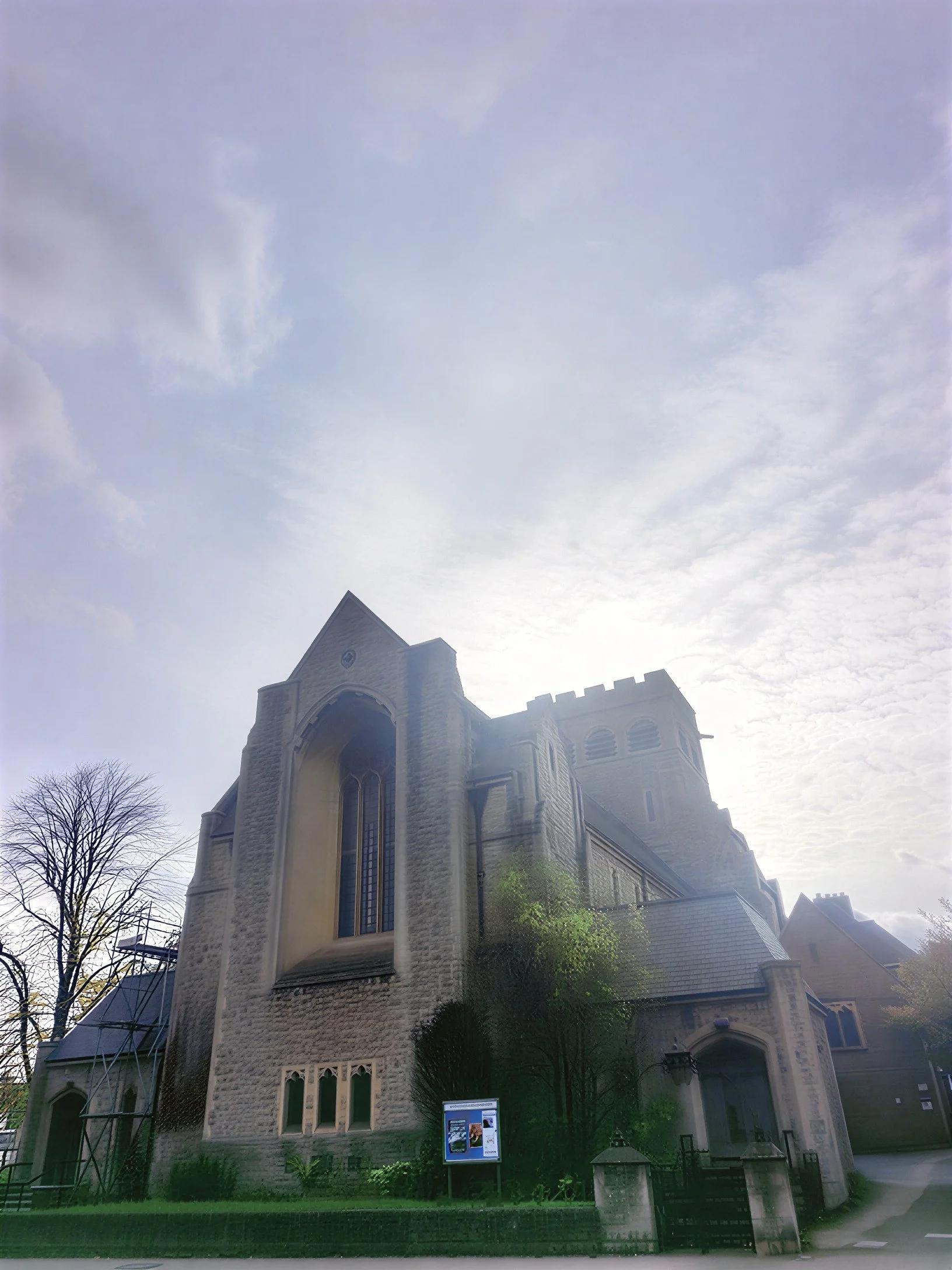 Penge Congregational Church. A large stone church with tall, narrow stained glass windows, surrounded by trees and a small garden, under a partly cloudy sky.