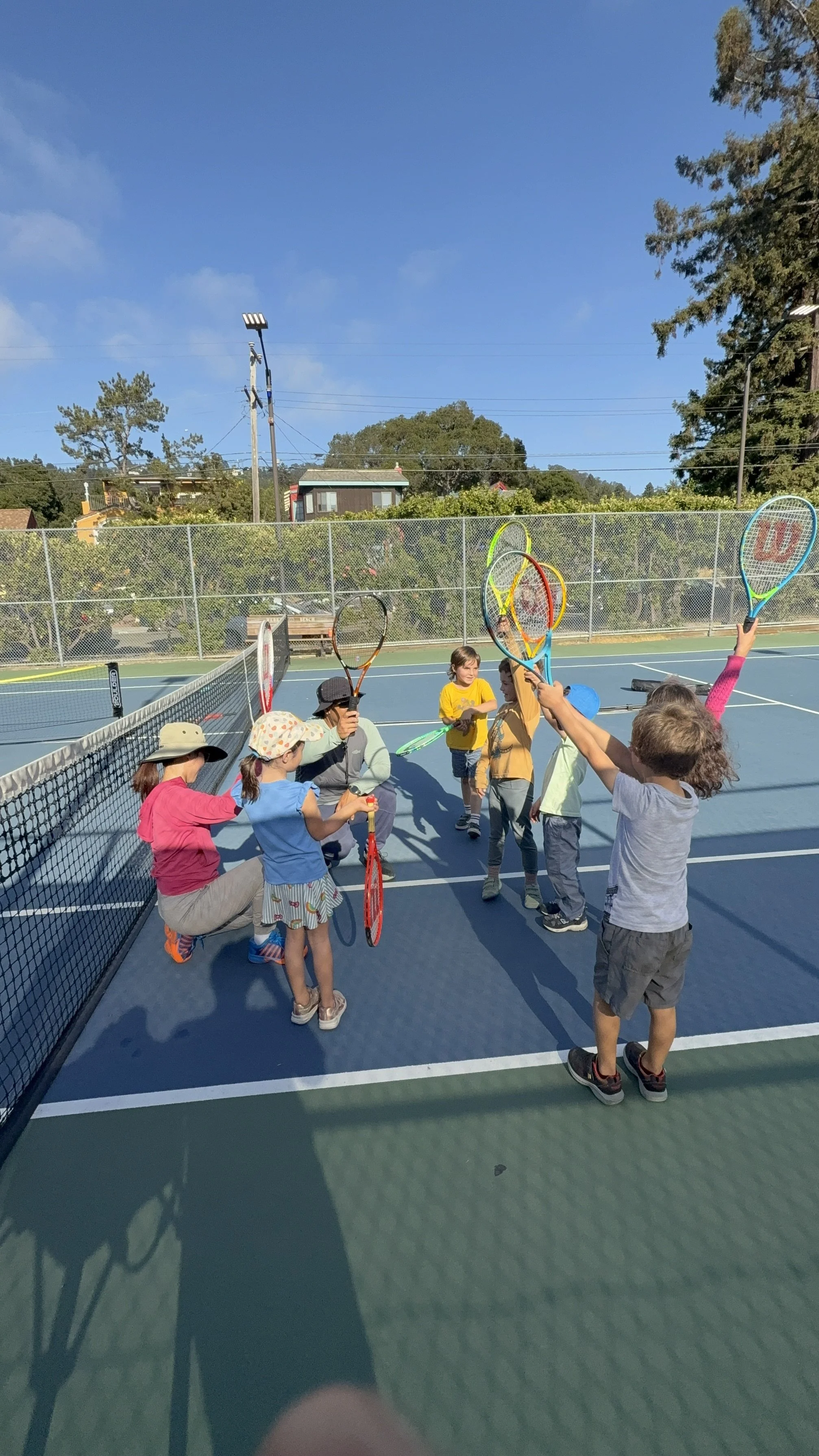 Children and a camp counselor on a tennis court, holding tennis rackets, some raising their rackets in the air, with a clear blue sky in the background.