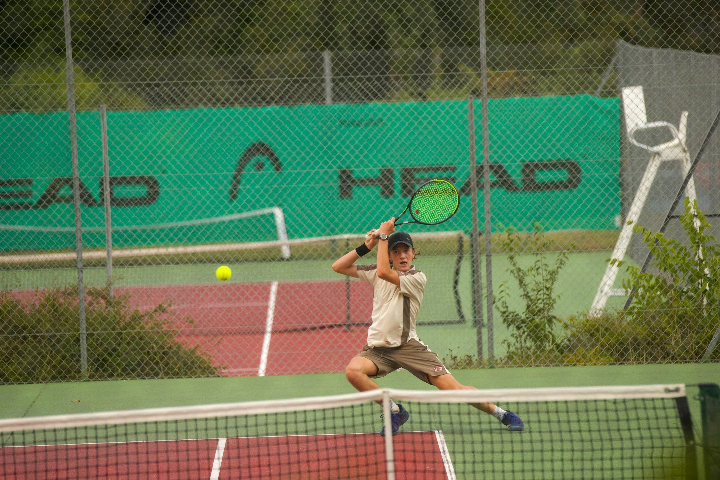 A young boy playing pickleball on an outdoor court, preparing to hit a yellow ball with a green paddle, with a net in the foreground and chain-link fence in the background.