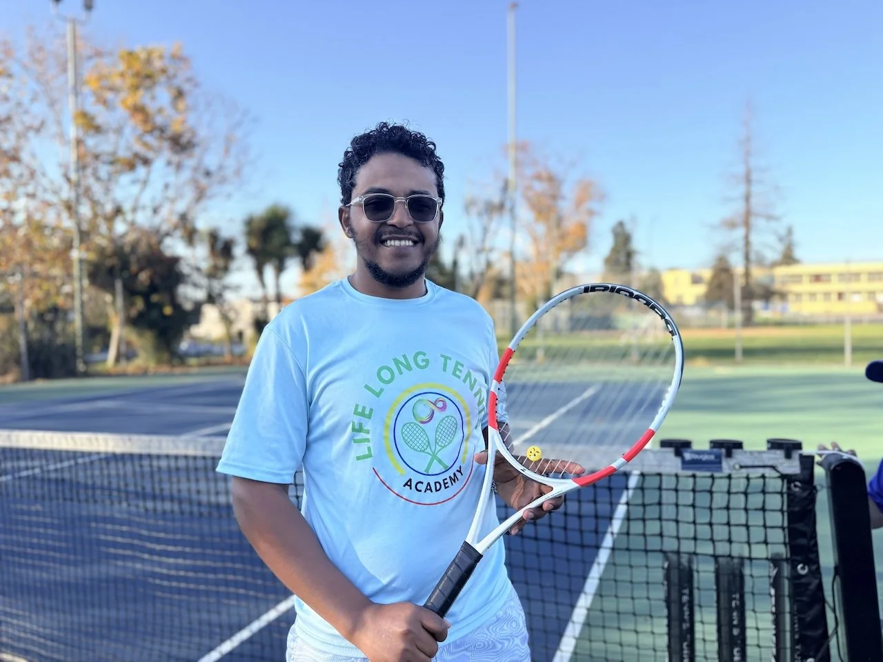 A man smiling on a tennis court holding a tennis racket with a tennis ball, wearing sunglasses and a light blue t-shirt with the logo 'Life Long Tennis Academy.'