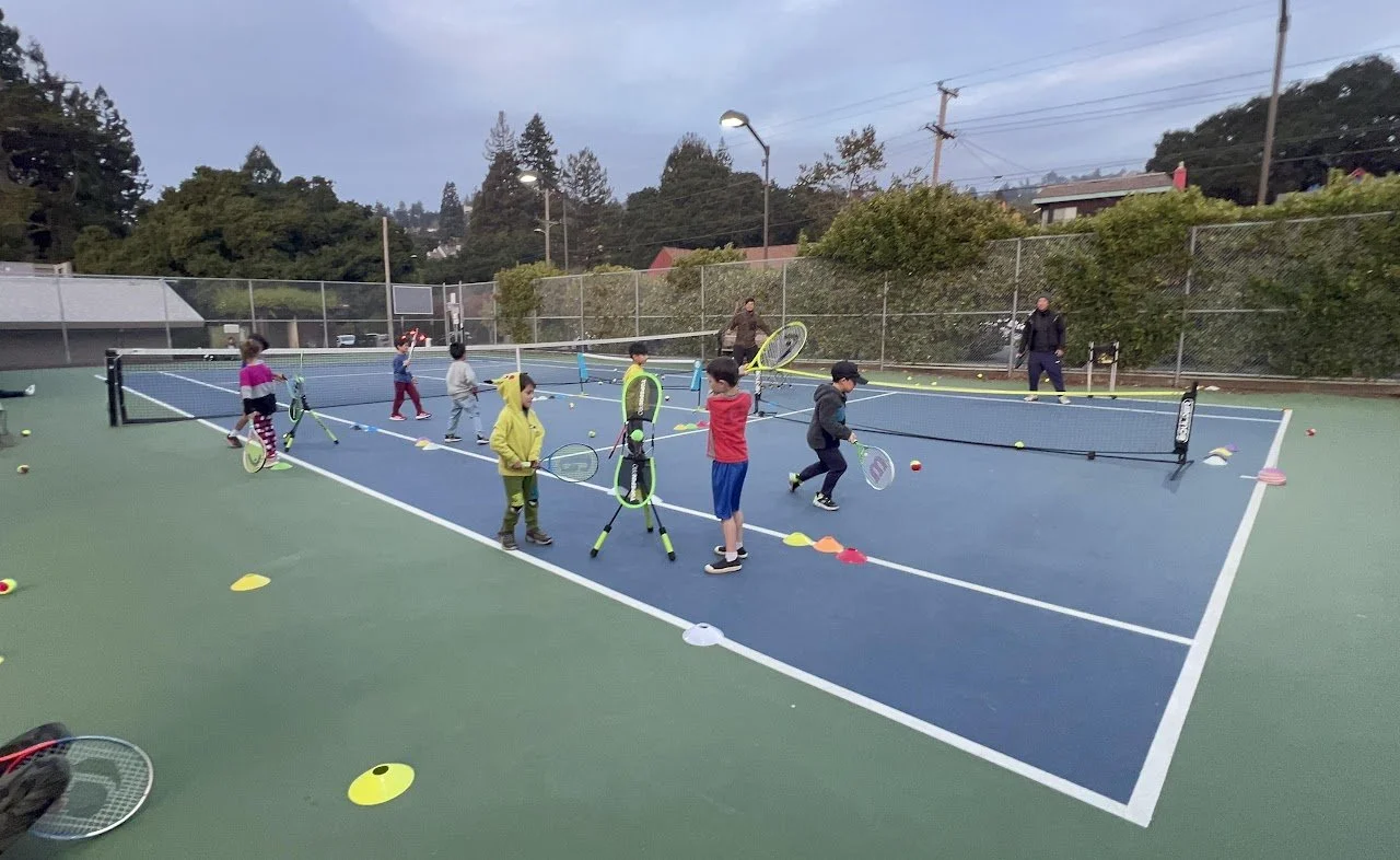 Kids playing tennis on an outdoor court with coaches, using tennis rackets and balls.