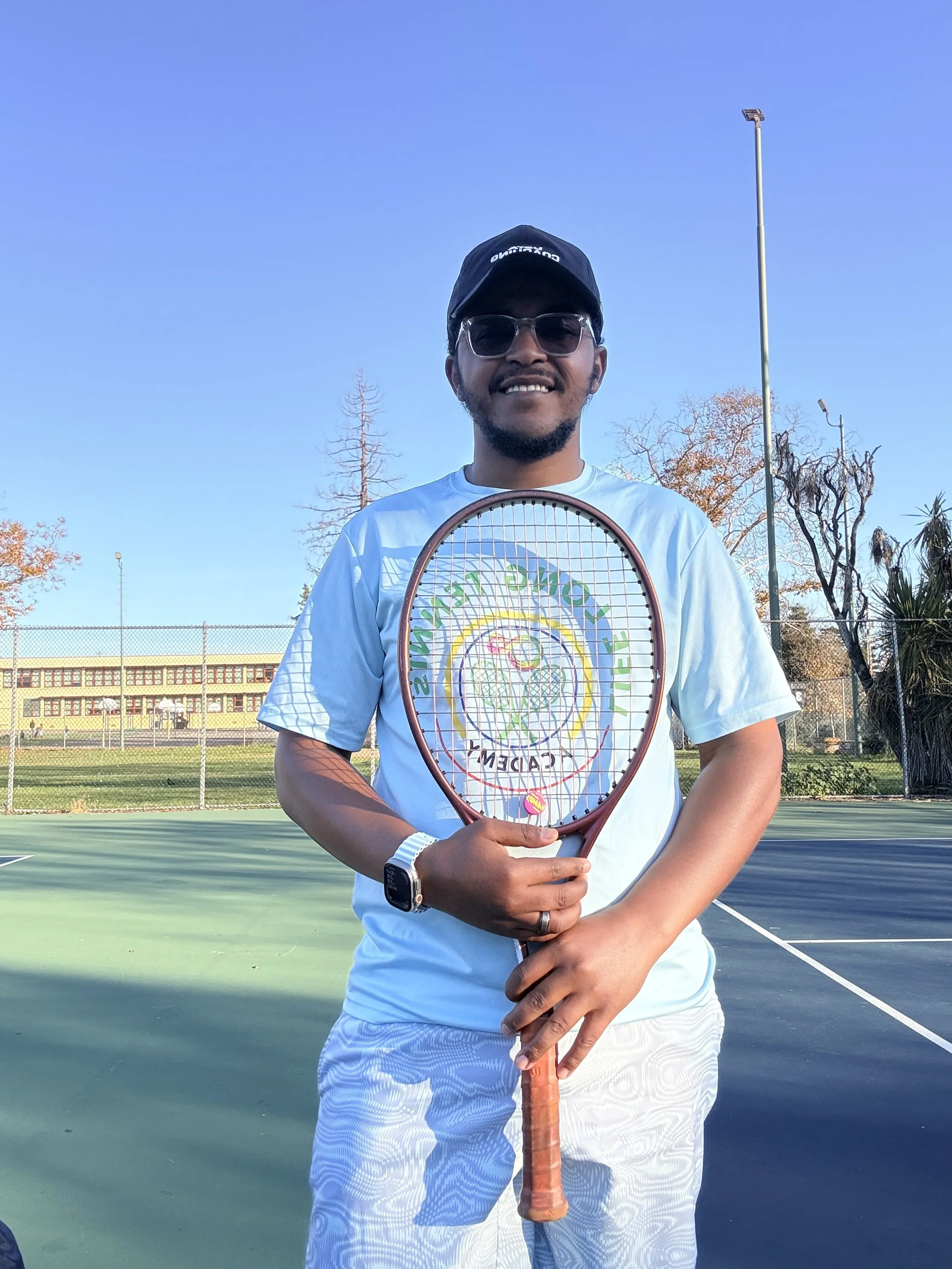 A man standing on a tennis court holding a tennis racket, wearing sunglasses, a black cap, a white sports shirt, and white patterned shorts, with a clear blue sky and tennis court fencing in the background.