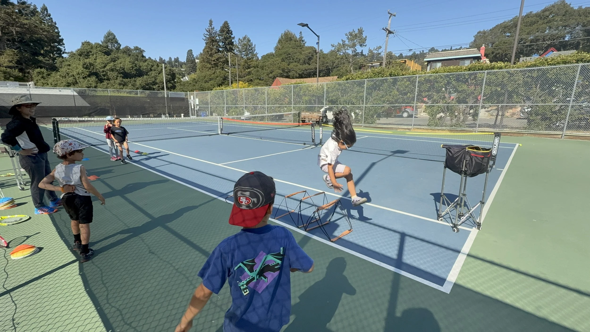 Children participating in a tennis drill on an outdoor tennis court with a coach overseeing the activity, surrounded by trees and residential houses under a clear blue sky.
