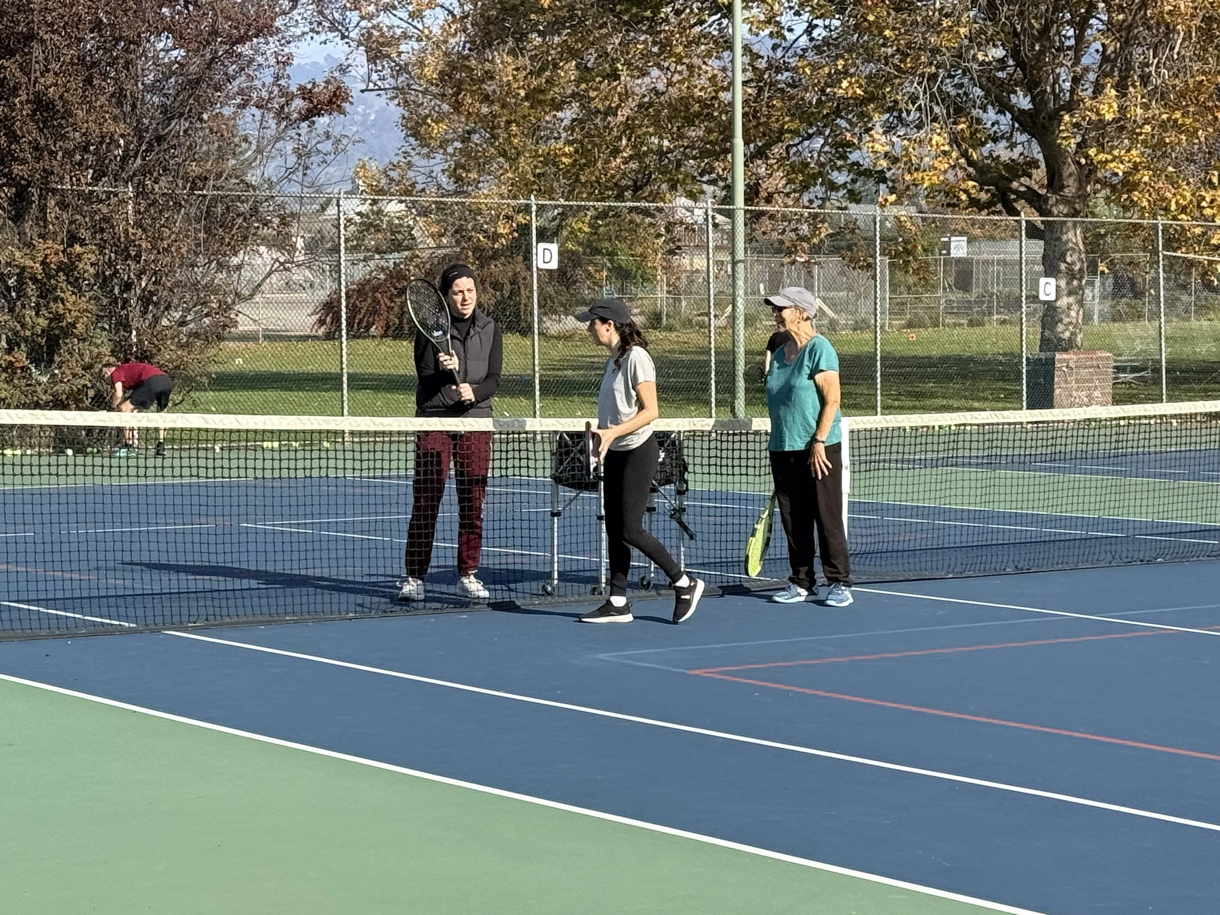 Three women standing on a tennis court near the net, holding tennis rackets, with one woman having a tennis cart nearby. A person in the background is picking up a tennis ball. Trees with autumn foliage are visible behind the fence.