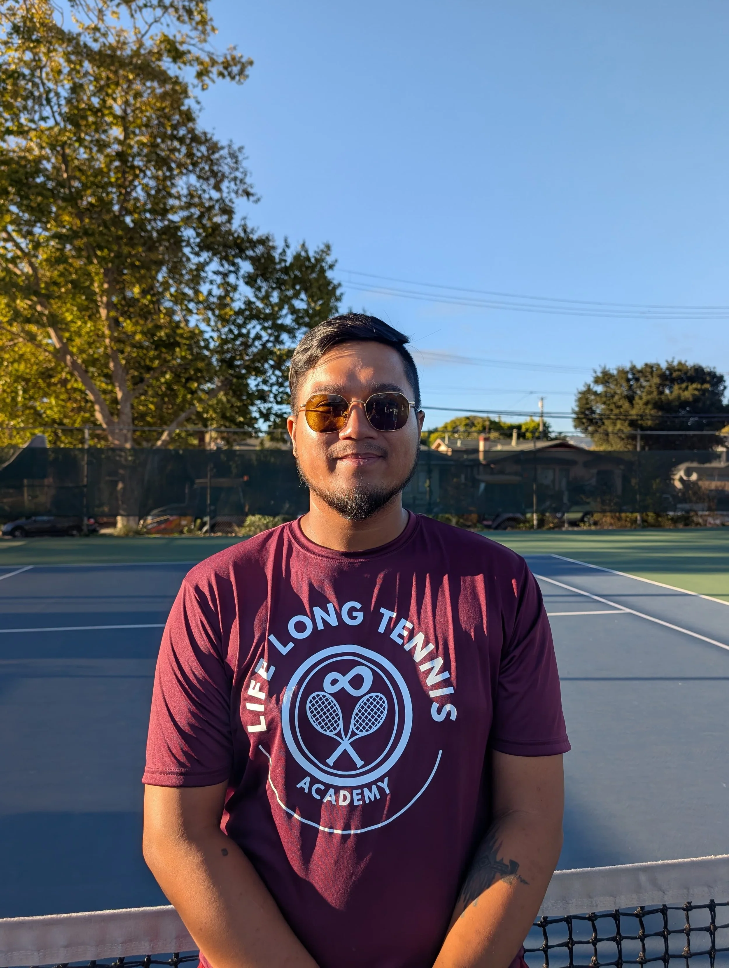 A man standing on a tennis court wearing sunglasses and a maroon T-shirt that reads 'Life Long Tennis Academy' with tennis rackets graphic.