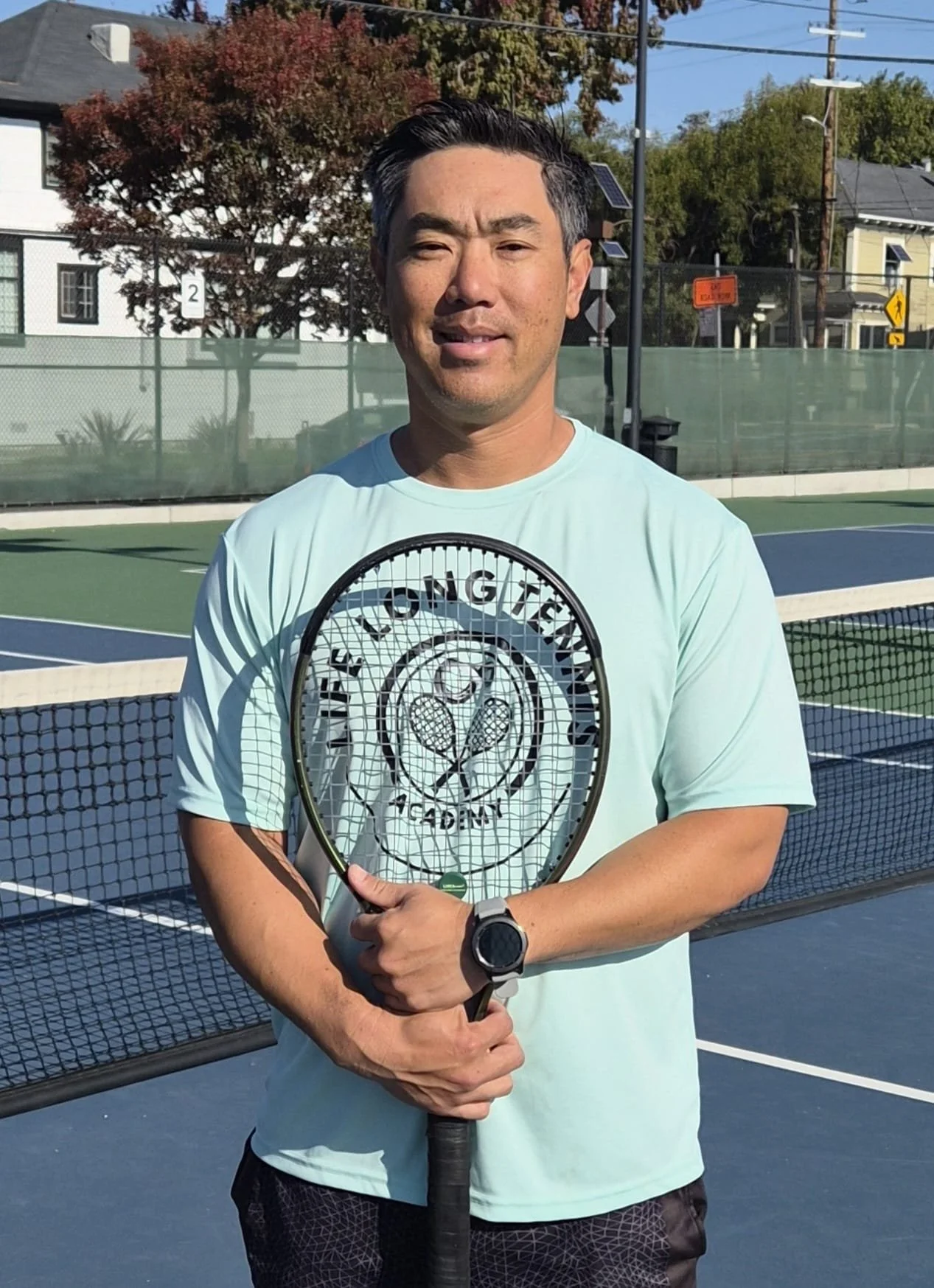 Man standing on a tennis court holding a tennis racket, wearing a light blue T-shirt, with trees, houses, and a blue sky in the background.