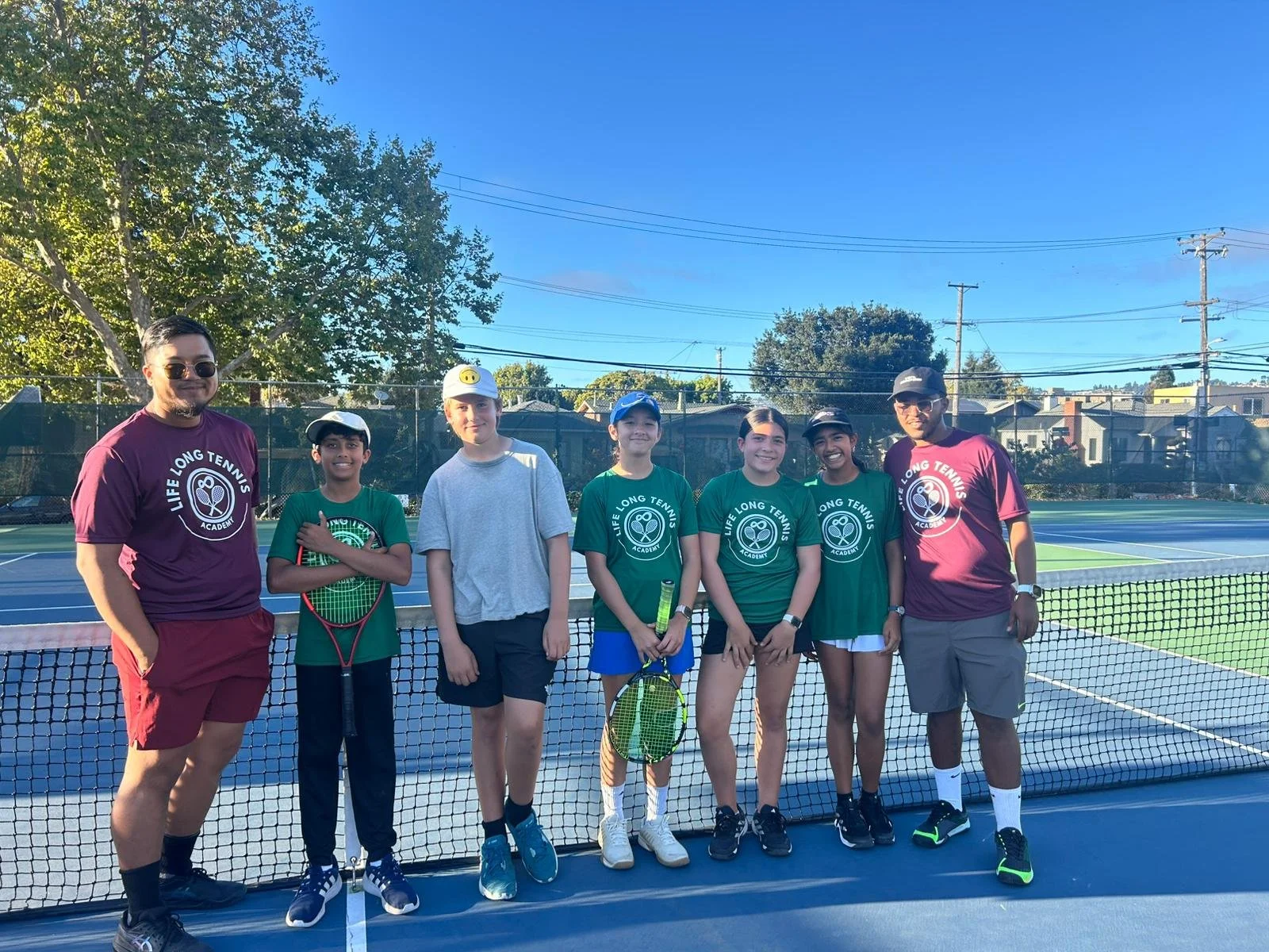 Seven people standing on a tennis court with a net in front of them, blue sky, trees, and power lines in the background. Four children wearing green shirts with tennis logos and two adults in maroon shirts with tennis logos. The children are holding tennis rackets.