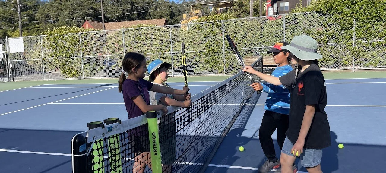 Children on a tennis court exchanging rackets during a learning session, with tennis balls on the ground, surrounded by trees and a chain-link fence.