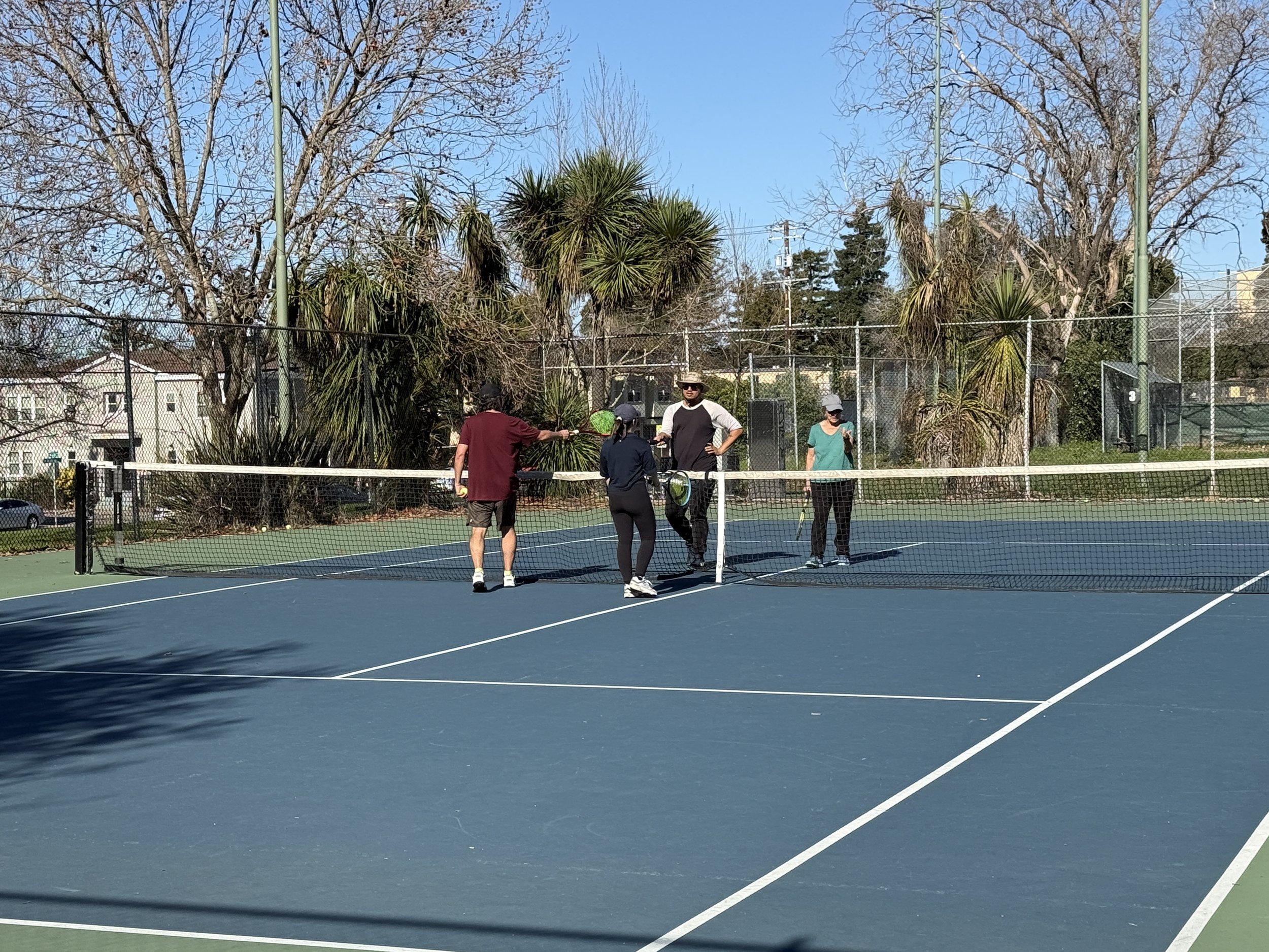 Four people standing on a tennis court, talking and taking a break. The court is outdoors with trees and houses in the background on a sunny day.