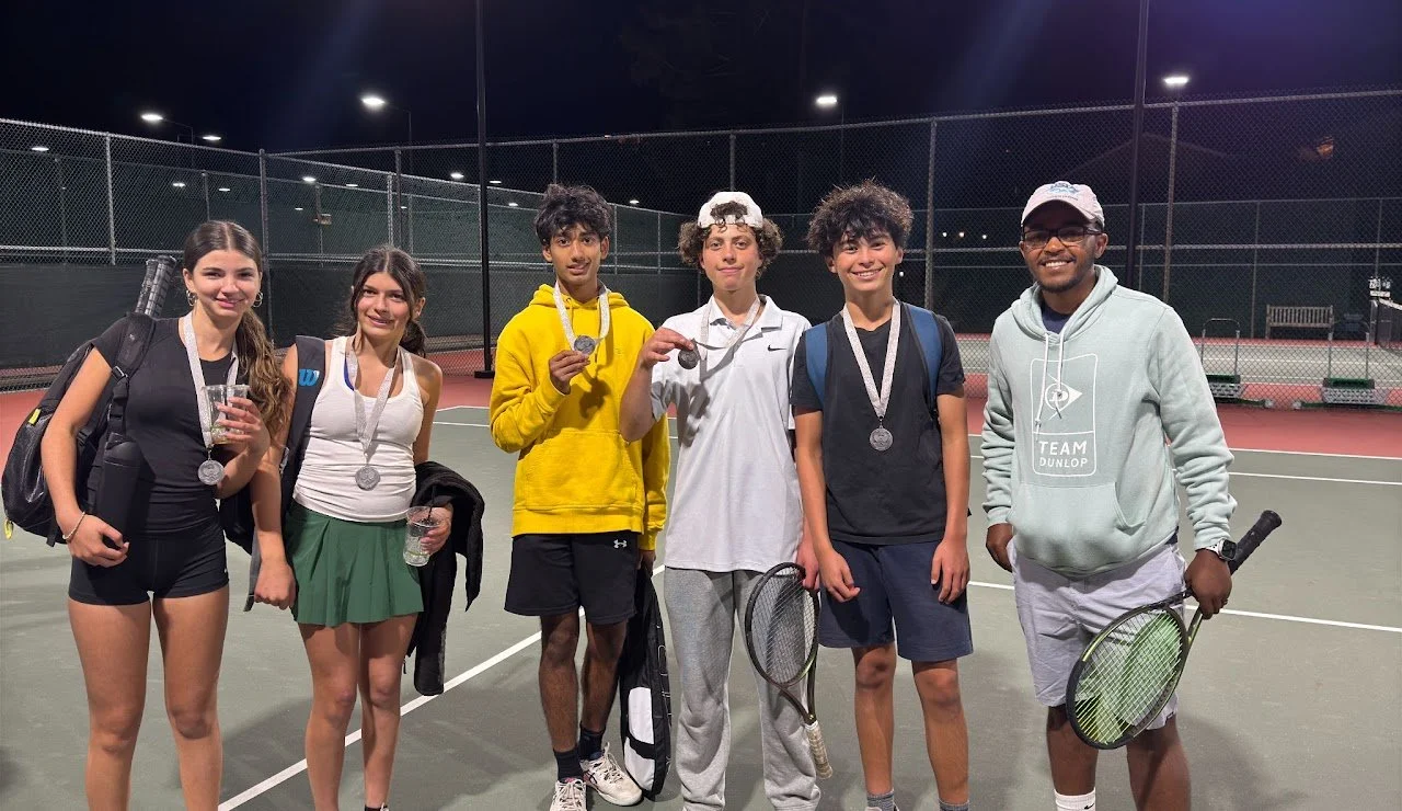 Group of six young tennis players standing on a tennis court at night, wearing medals, holding tennis rackets, and smiling.