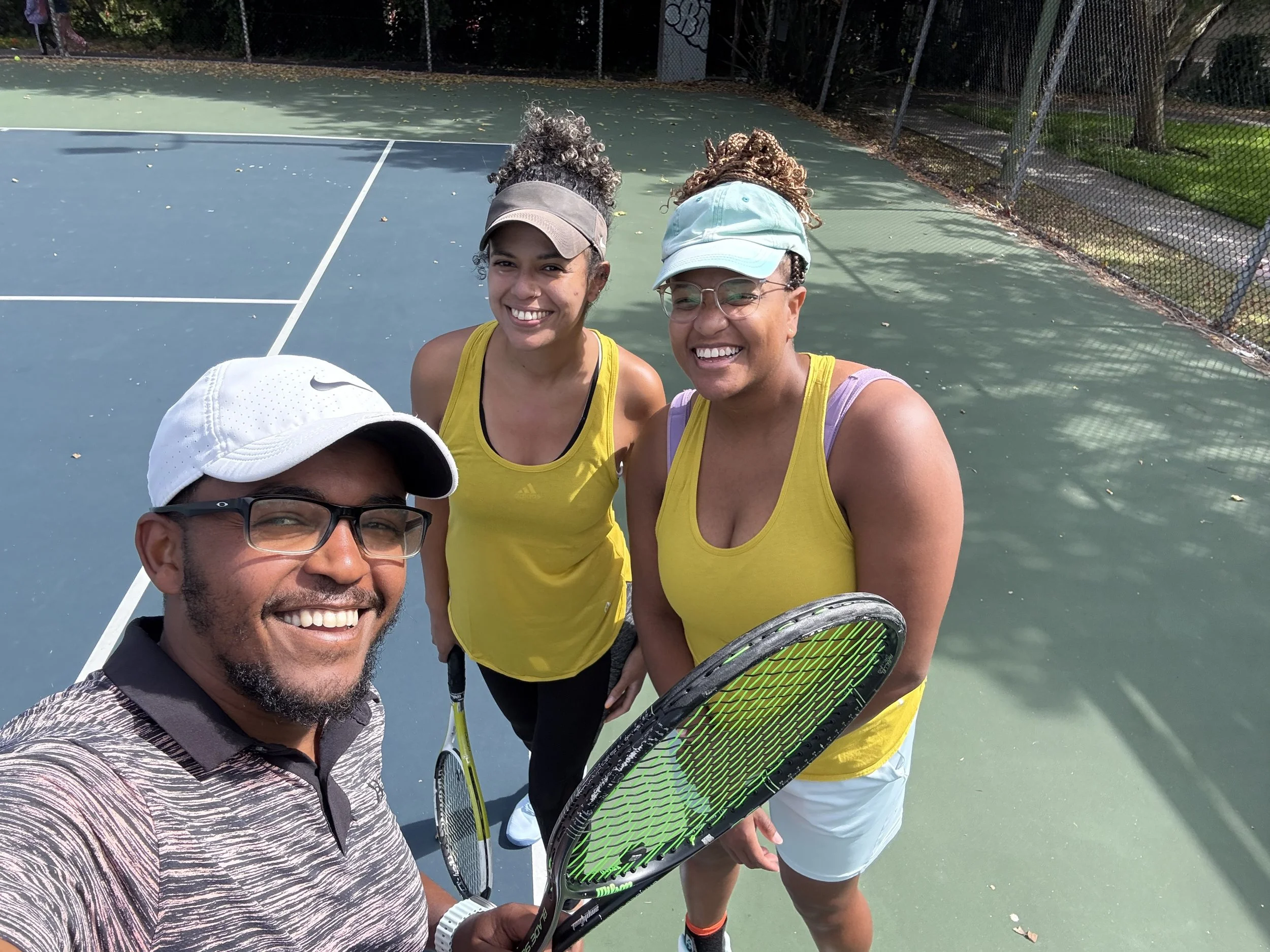 Three people smiling on a tennis court, holding tennis rackets, dressed in athletic wear.