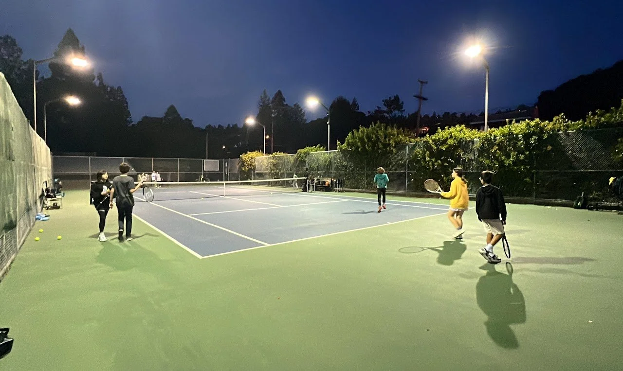 People playing tennis on an outdoor court at dusk, with some standing and some actively hitting the ball.