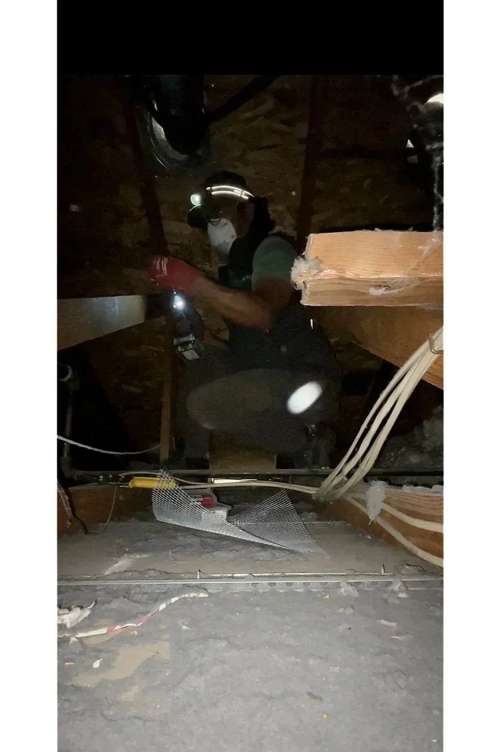 A person working in a dark attic or ceiling space, using a headlamp, surrounded by exposed wooden beams and electrical wiring.