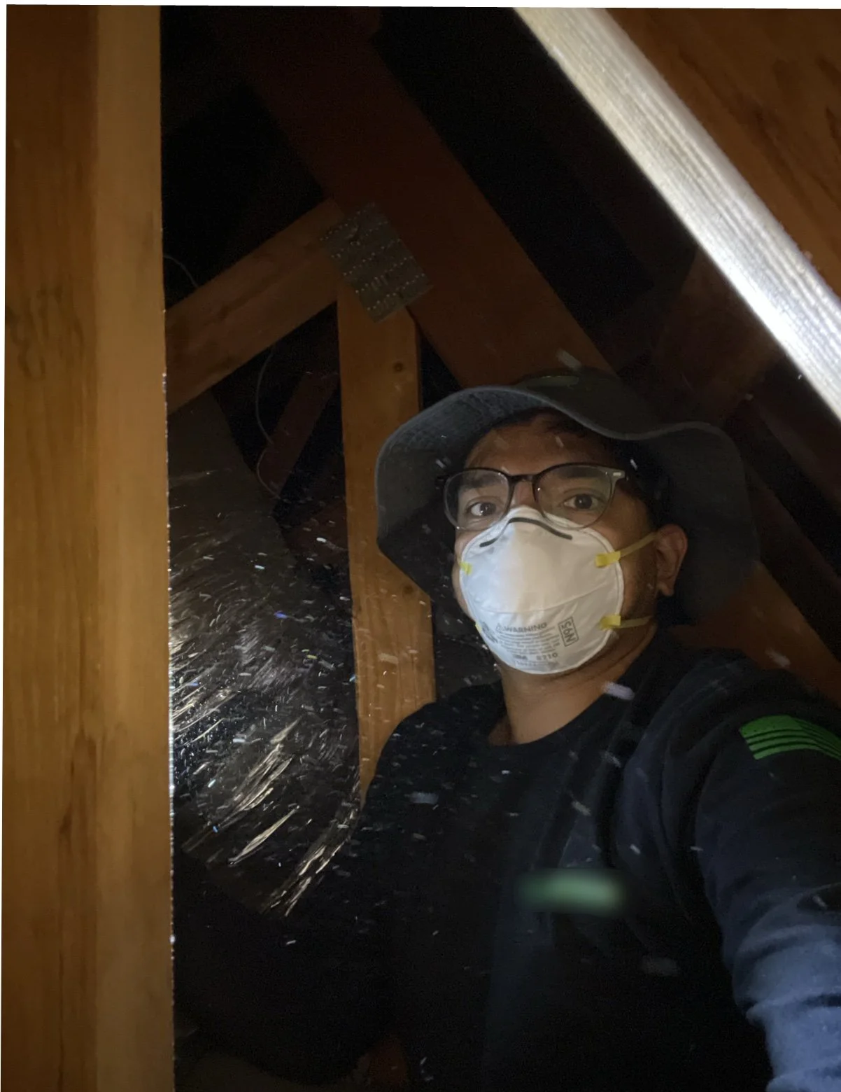 A person wearing a hat, glasses, a face mask, and a black jacket taking a selfie inside an attic with wooden beams and insulation.