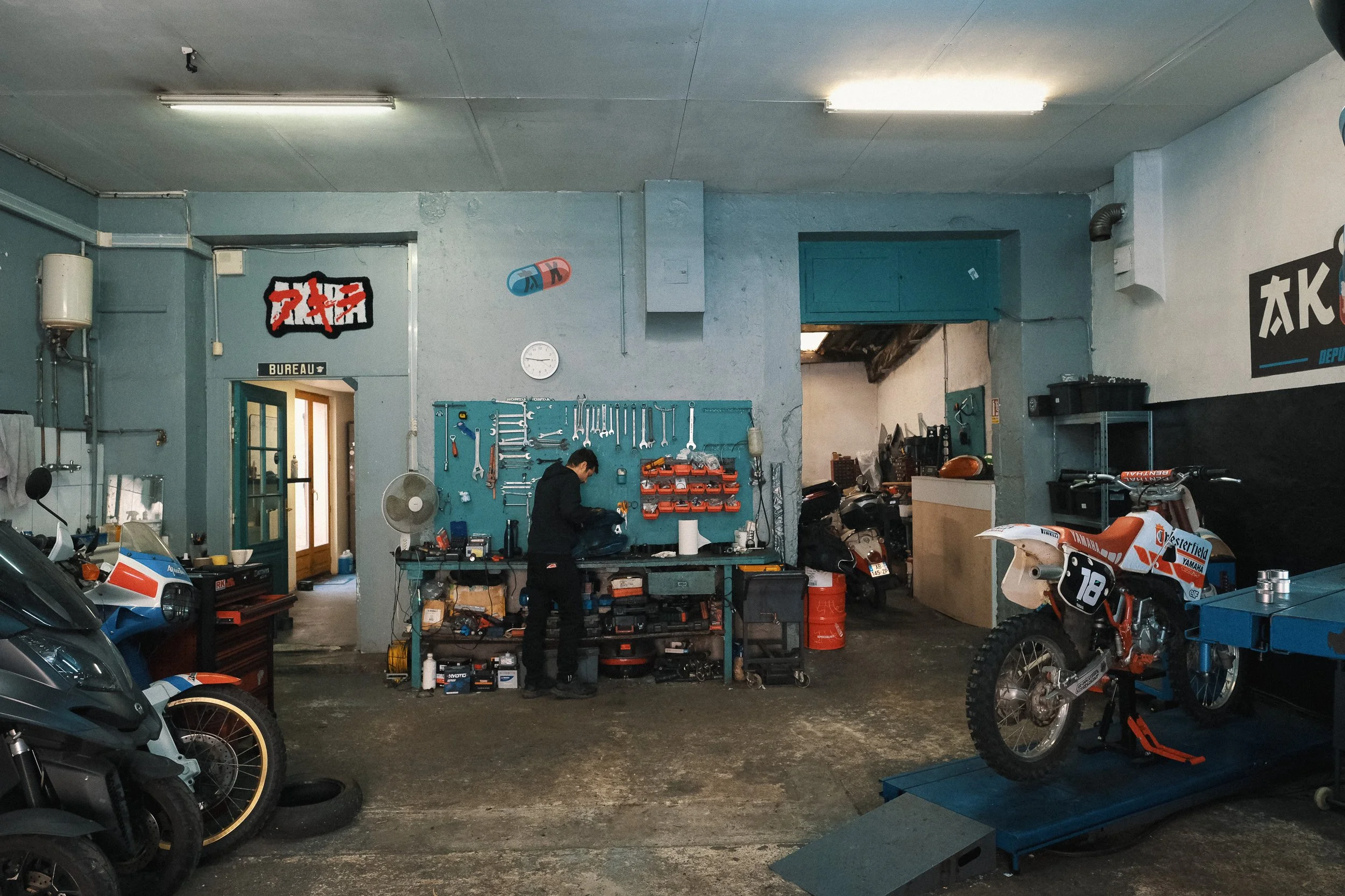 Atelier de réparation de motos avec un homme travaillant à une table, motos en attente de réparation, outils suspendus au mur, et un mur peint en bleu et blanc.