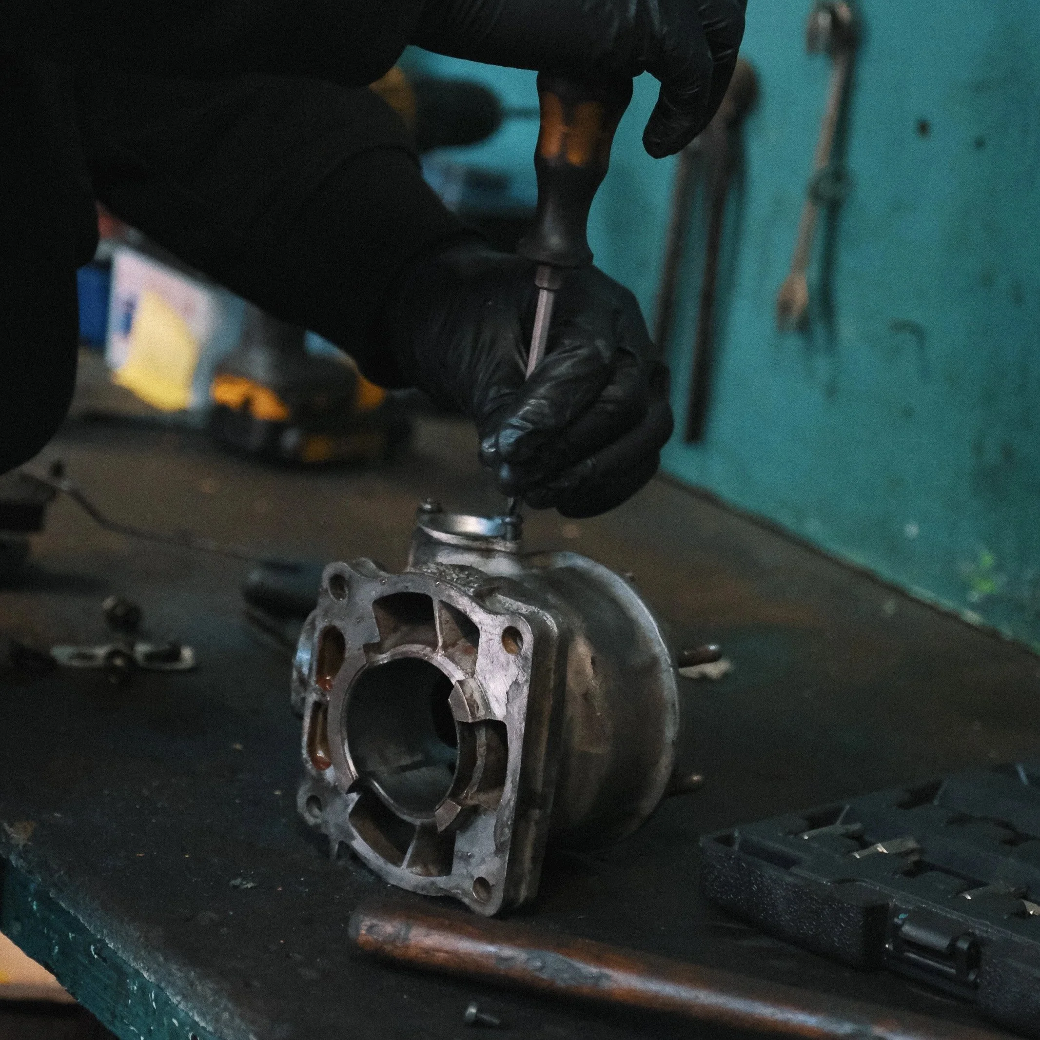 Une personne portant des gants noirs travaille sur une pièce mécanique à l'aide d'un tournevis dans un atelier de réparation automobile.