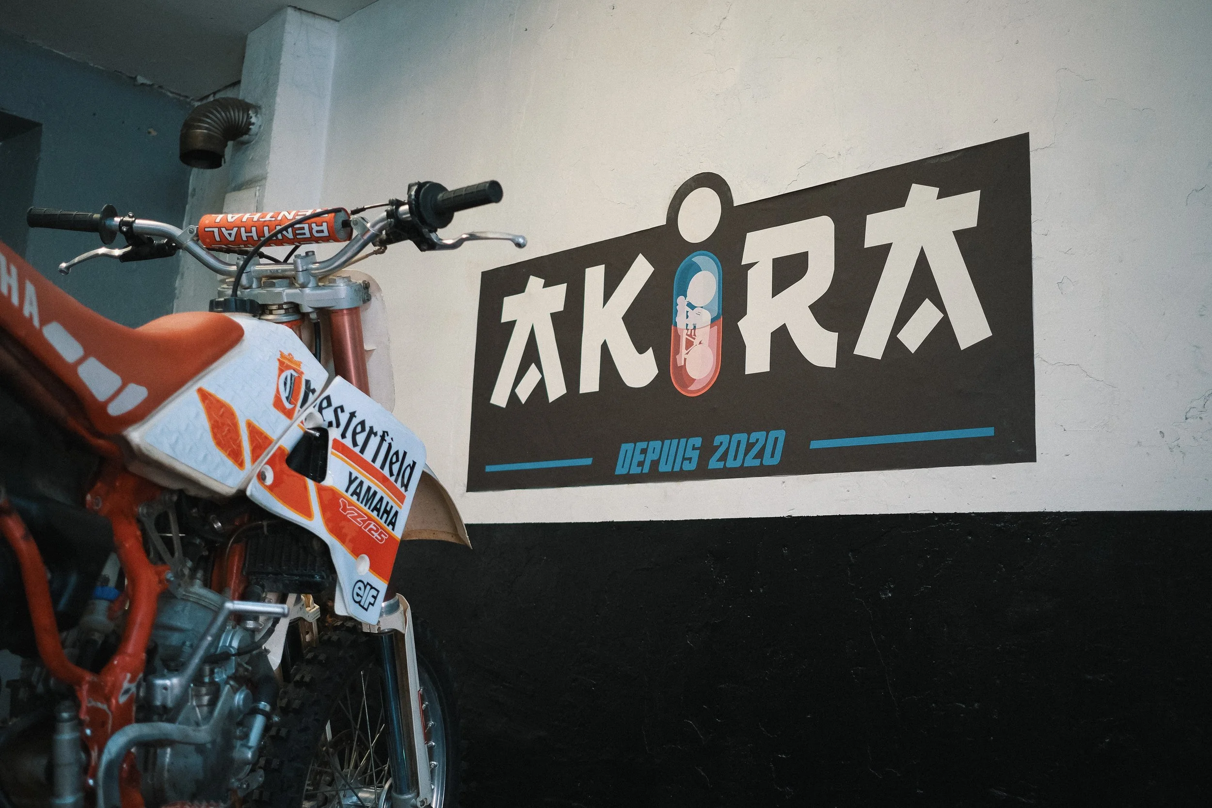 Une moto tout terrain orange et blanche avec des logos Yamaha, Elf, et Westernfield, stationnée devant un mur blanc avec un panneau promotionnel de l'entreprise Akora, fond noir, indiquant 'Depuis 2020'.
