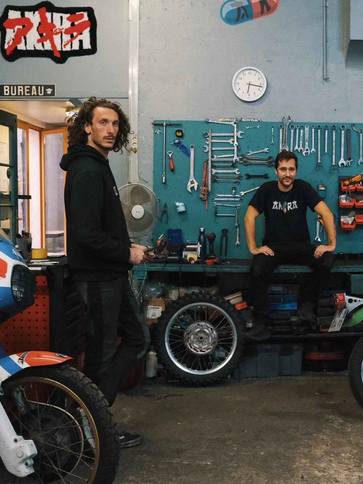 Deux hommes dans un atelier de réparation de motos, l'un debout et l'autre assis sur une table de travail. L'atelier a des outils suspendus sur un panneau bleu derrière eux, un mur avec une horloge, et une moto partiellement visible en premier plan.