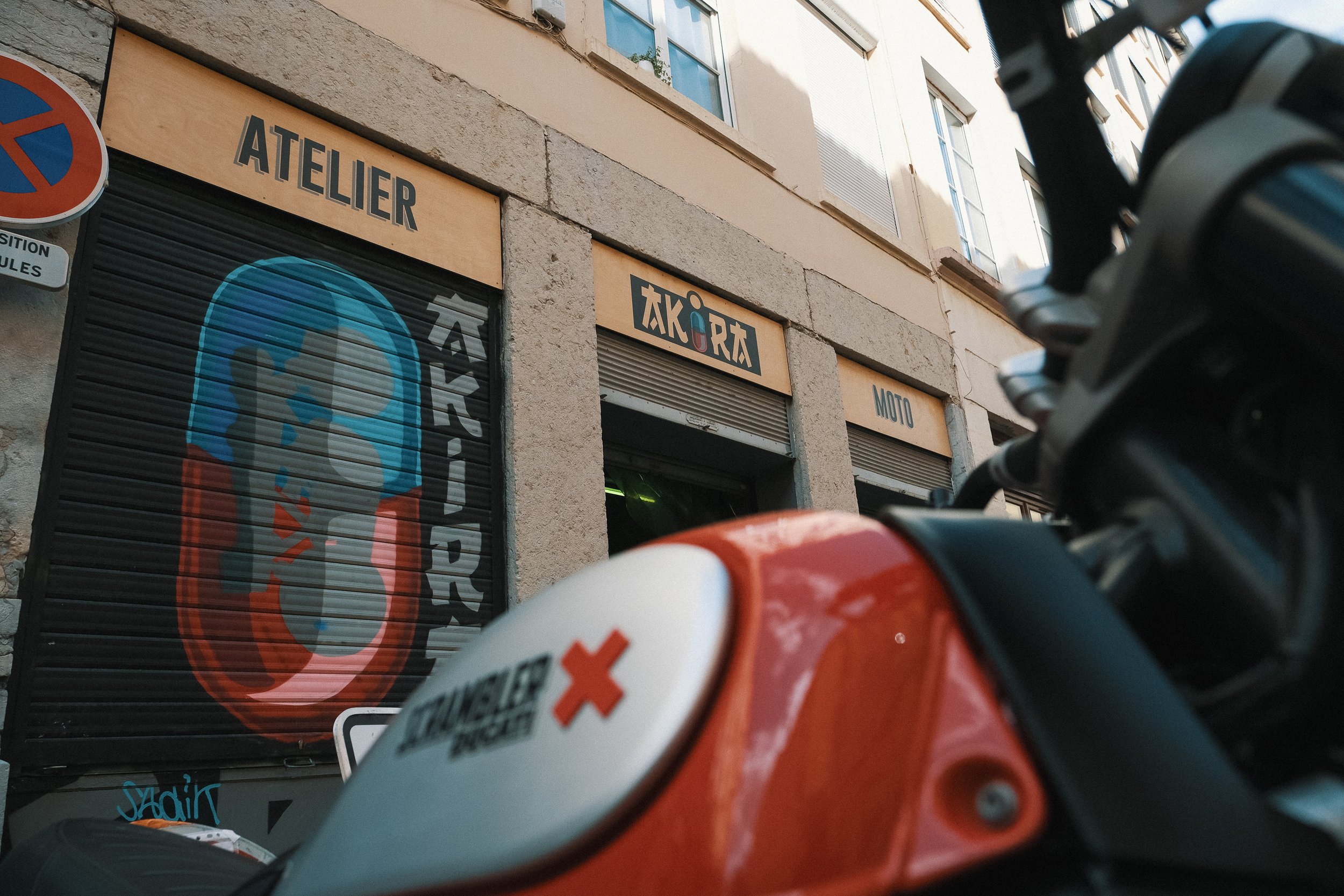 Façade d'un atelier de réparation moto avec un logo représentant un casque de motard, peint sur une grille métallique et intitulé 'ATELIER TAKORA MOTO' en lettres bleues sur une enseigne en bois, avec un scooter orange en premier plan.