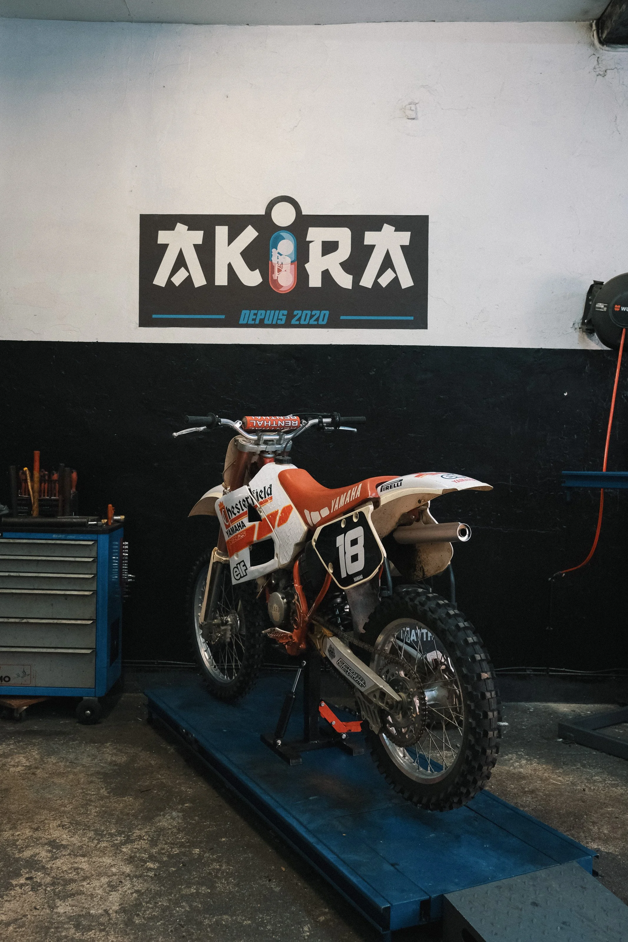 Une moto de motocross Yamaha sur un stand dans un garage, avec un mur noir et blanc derrière et un logo 'AKIRA' avec la mention 'Depuis 2020' peint sur le mur.
Garage Akira motors entretient et réparation moto à Lyon.
