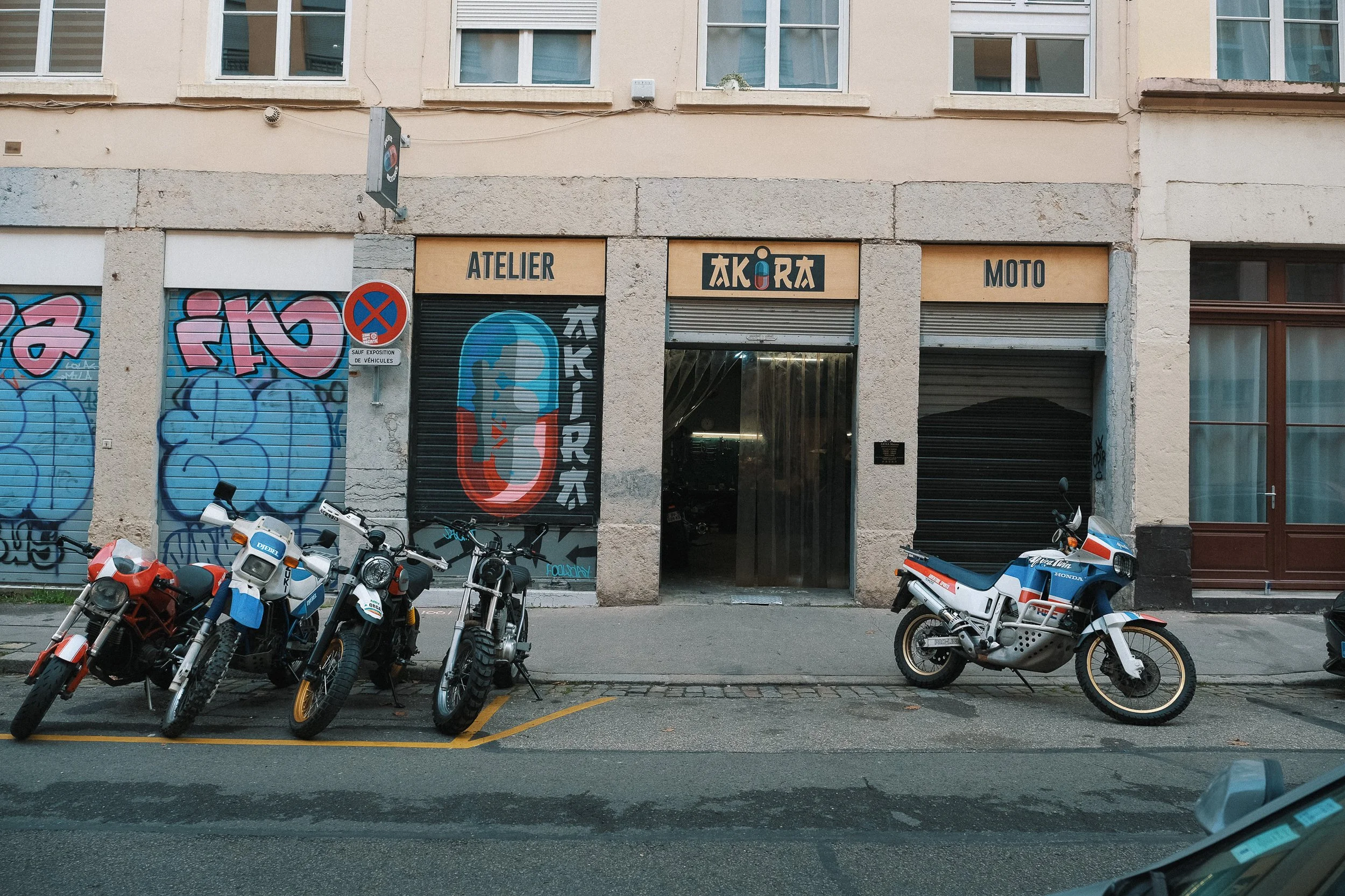 Façade d'un bâtiment avec cinq motos garés devant, graffitis sur les volets roulants, enseignes indiquant 'Atelier Akira Motors'.  Garage Akira motors entretient et réparation moto à Lyon.