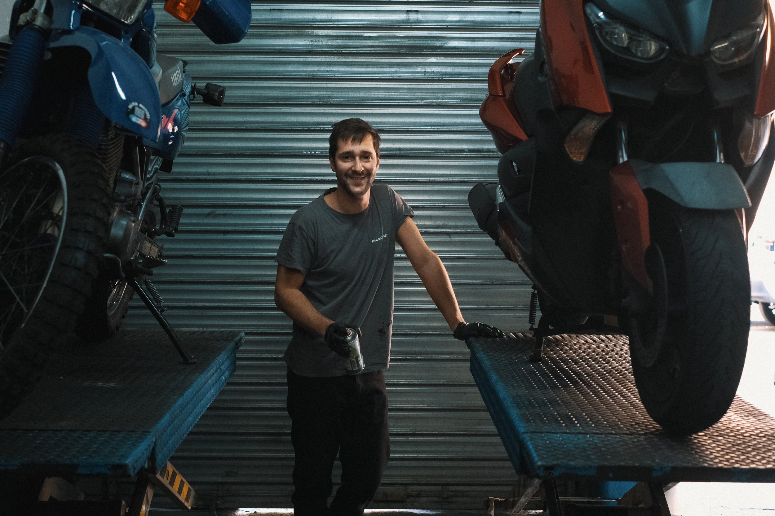 Un homme souriant, portant des gants noirs, se tient entre deux motos ou scooters élevés sur des plateformes dans un garage, avec une porte roulante métallique en arrière-plan.
 Garage Akira motors entretient et réparation moto à Lyon.