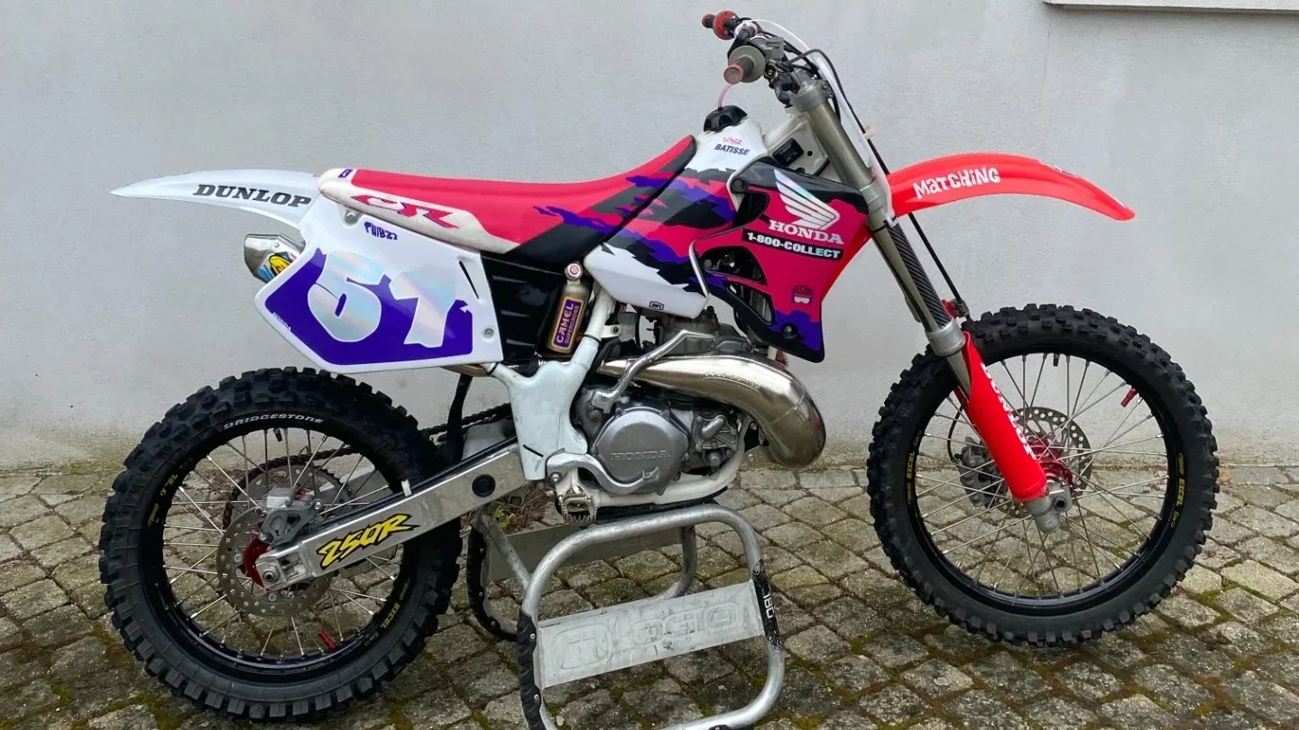 Moto tout-terrain de course avec un cadre rouge, des pneus à crampons, et des sponsors comme Dunlop, Honda, et Matching, stationnée sur un pavé face à un mur gris.  Garage Akira motors entretient et réparation moto à Lyon.