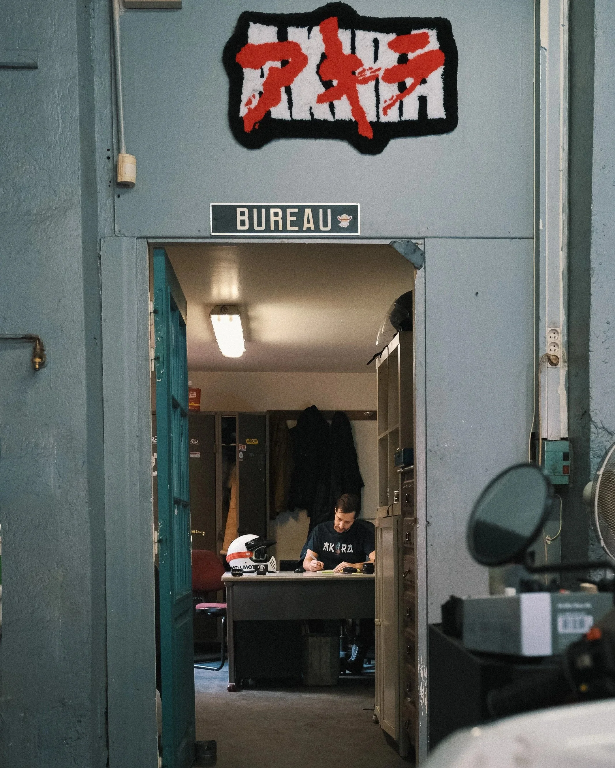 Vue d'une salle de bureau vue à travers une porte entrouverte, avec un homme assis à un bureau, portant un t-shirt noir et écrivant, un casque de moto blanc et noir sur le bureau, et des vestes accrochées au mur en arrière-plan.