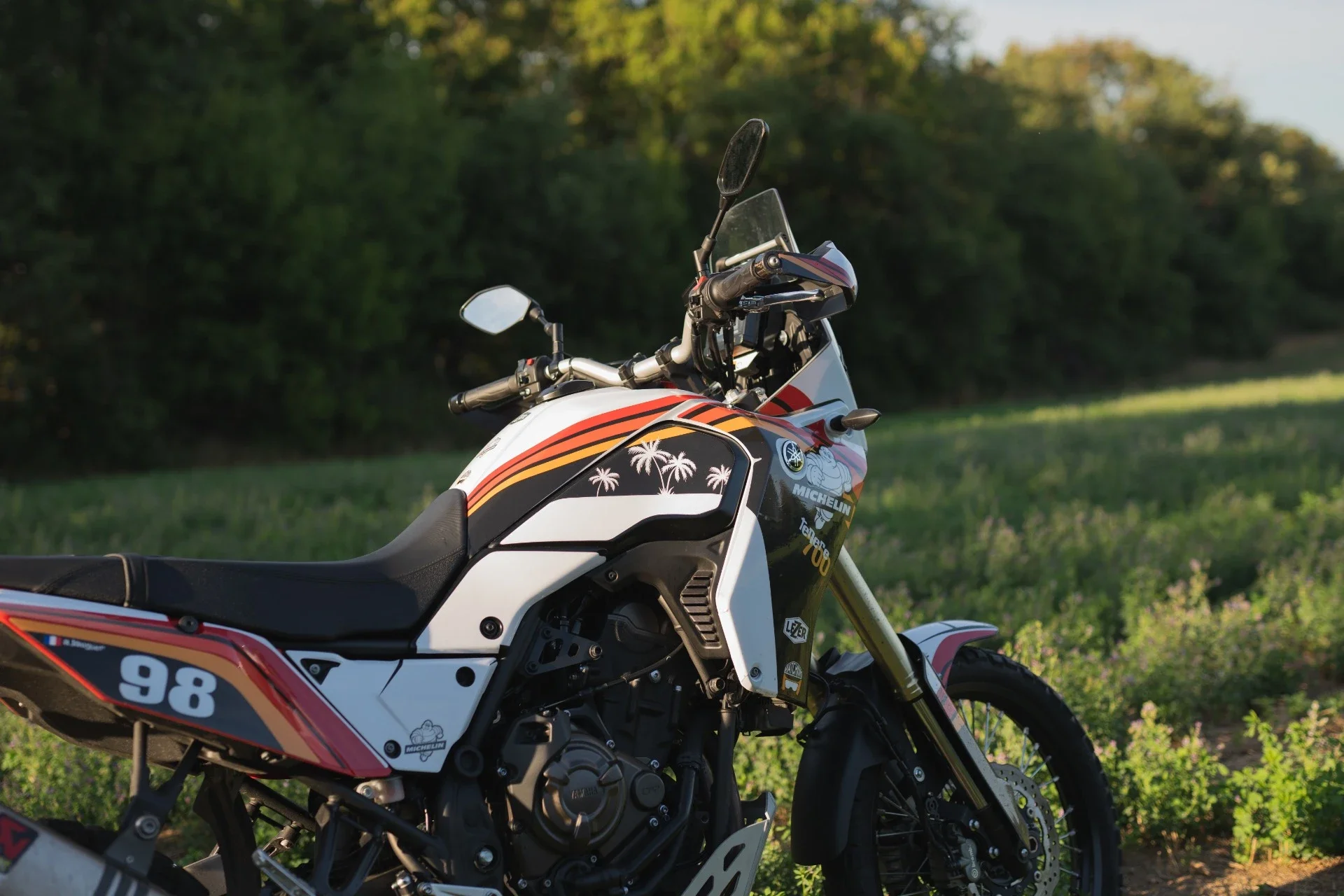 Moto de rallye Yamaha Ténéré 700 avec numéros 98, décorée de palmiers et logos Michelin, sur une route entourée de végétation.
Garage Akira motors entretient et réparation moto à Lyon.