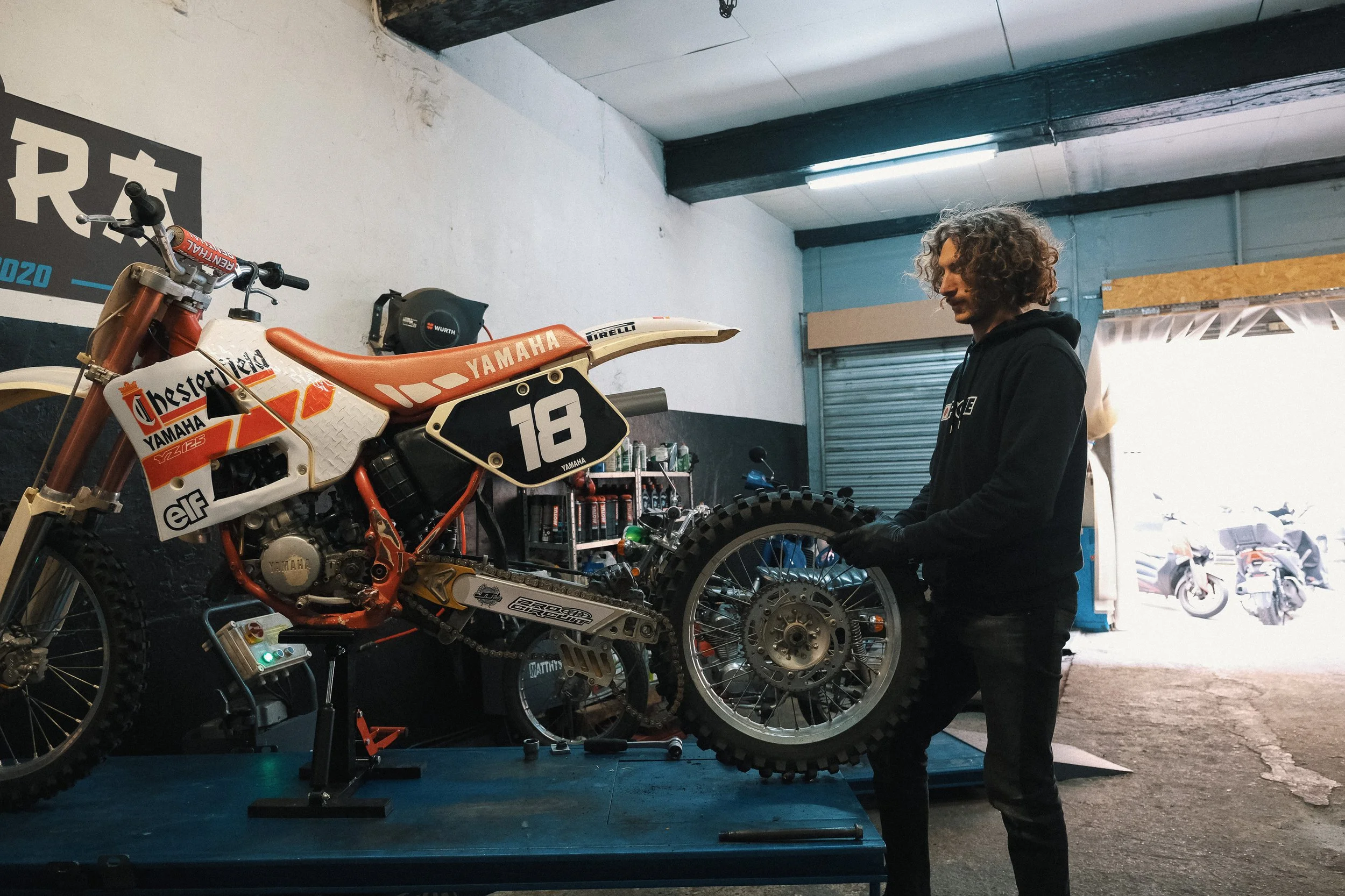 Un homme au garage réparant une moto de motocross Yamaha orange et blanche kit déco Chesterfield. Garage Akira motors entretient et réparation moto à Lyon.