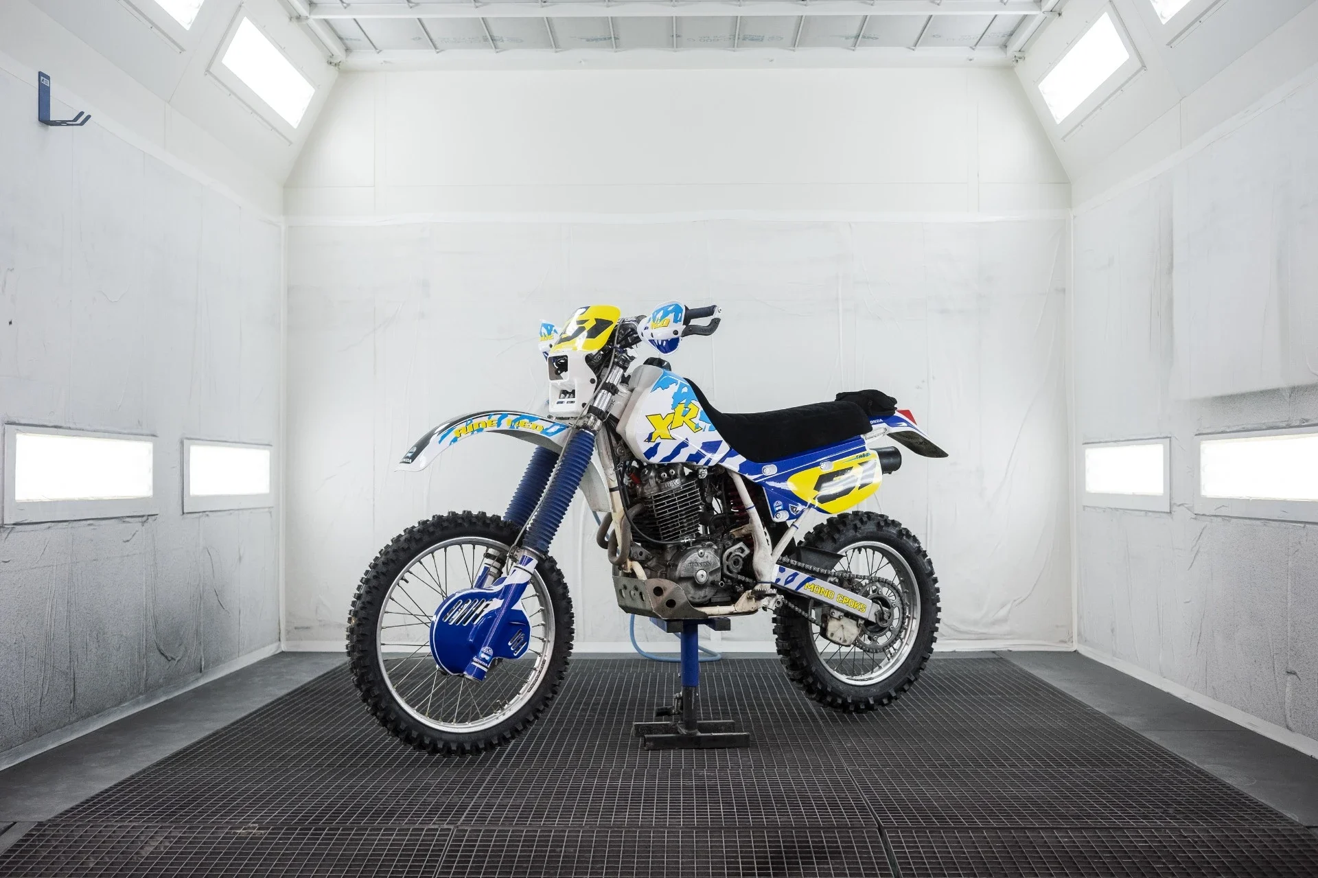 Motocross bleue, blanche, jaune avec protection et pneus crantés, dans une cabine d'exposition.
Garage Akira motors entretient et réparation moto à Lyon.