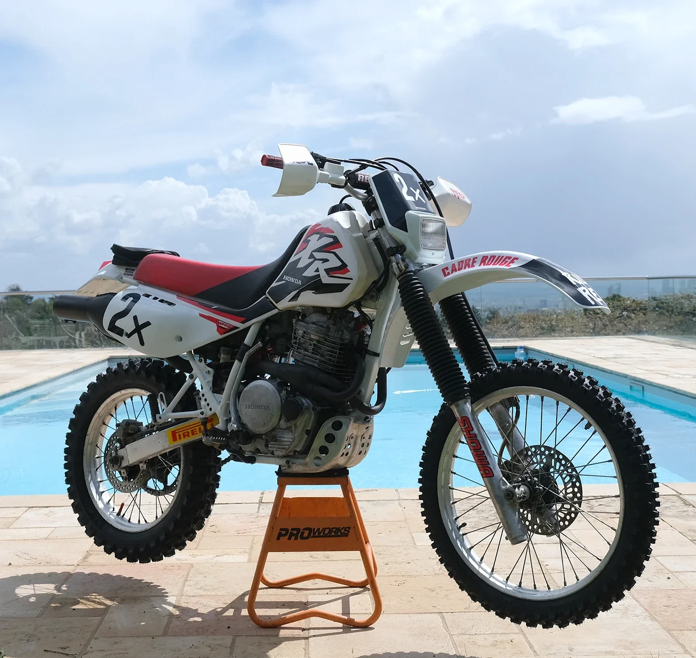 Une moto tout-terrain Honda 600 XR suspendue sur un support orange près de la piscine. Le fond montre un ciel nuageux et un extérieur avec vue panoramique.  Garage Akira motors entretient et réparation moto à Lyon.