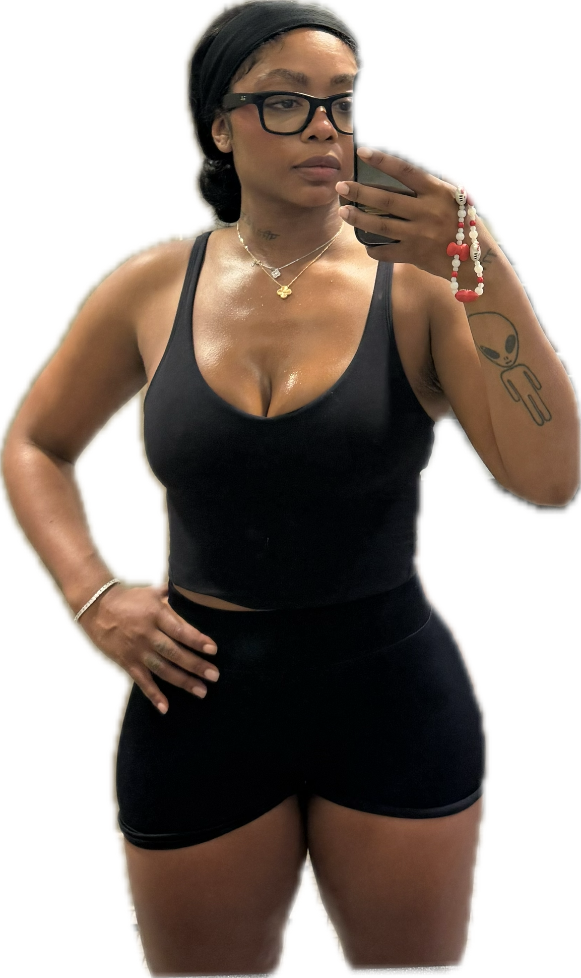 A woman taking a mirror selfie, wearing sunglasses, a black tank top, and black shorts, with tattoos on her left arm and a necklace, and holding a smartphone in her right hand.