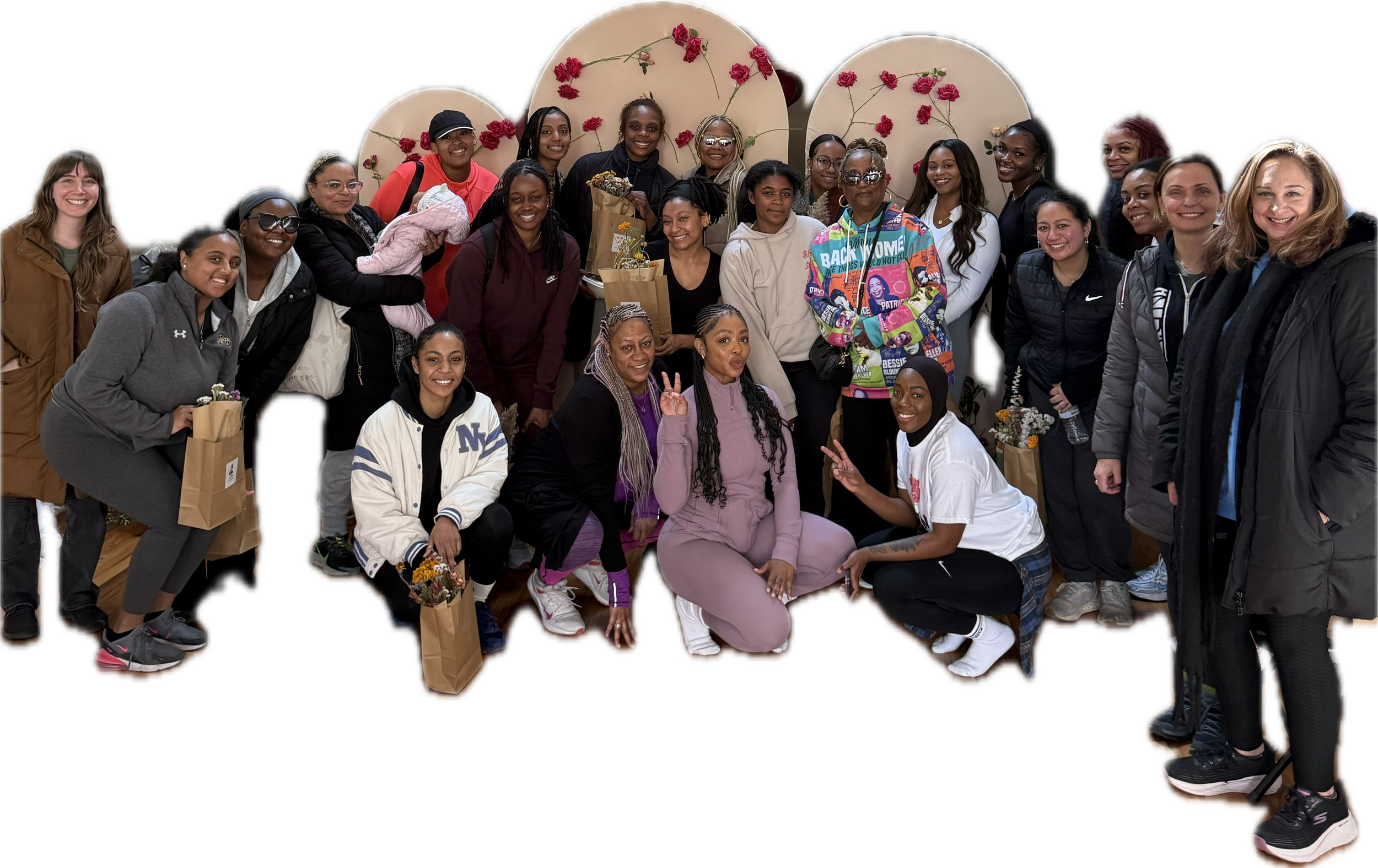 Group of diverse women gathered together, some holding flowers and making peace signs, in an indoor setting with decorative backdrop.