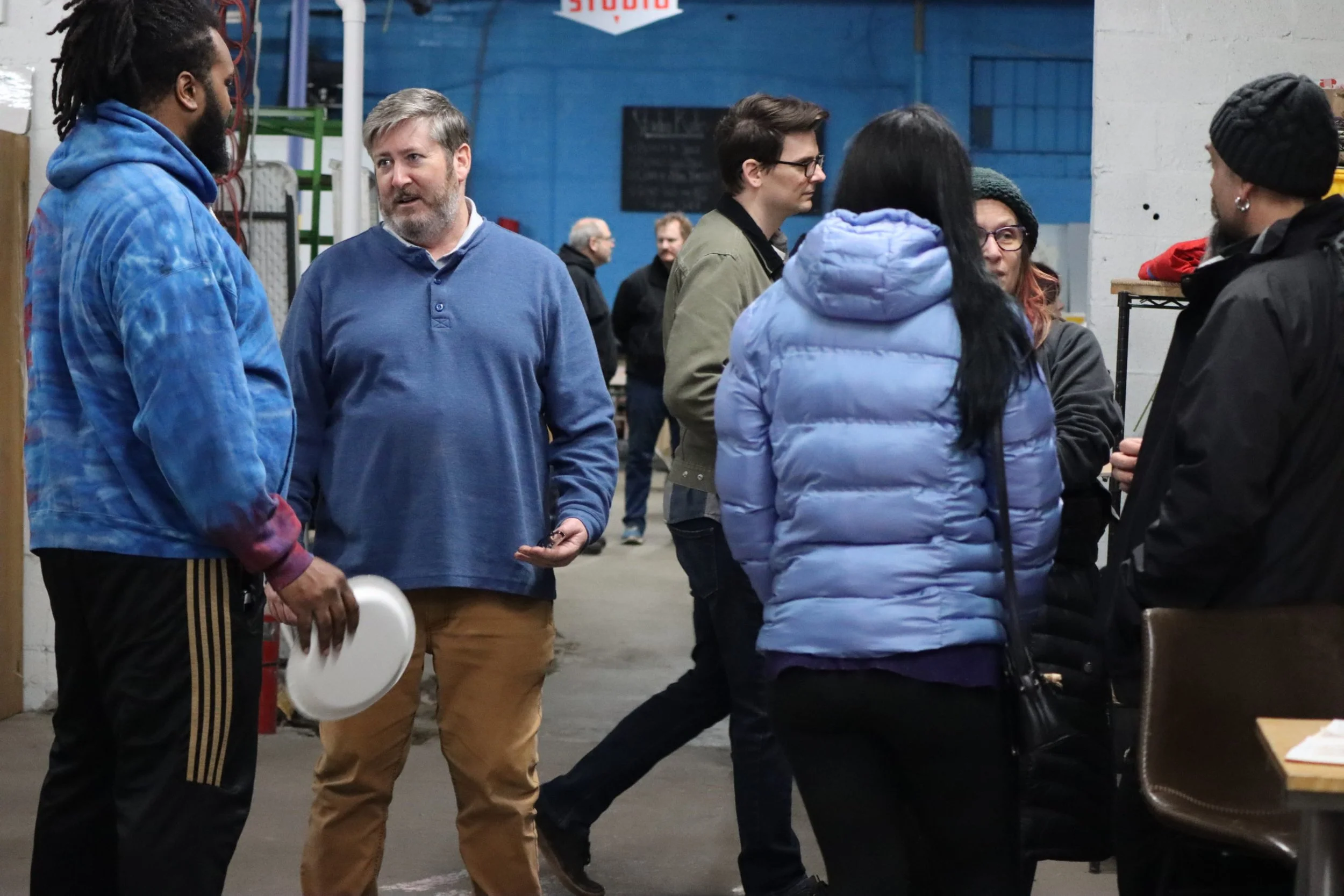 A group of six people standing and talking in a warehouse or industrial space, with some shelves and a blue wall in the background.