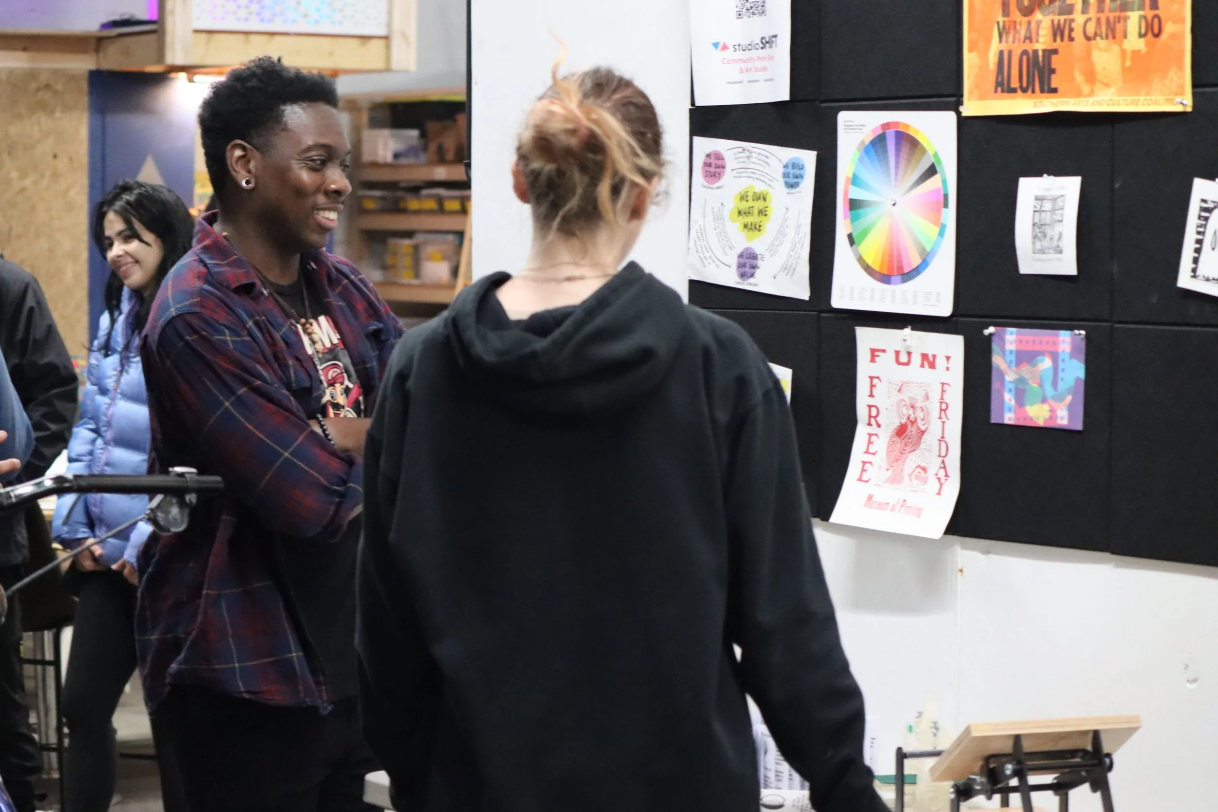 Two people talking in front of a black bulletin board decorated with colorful posters and artwork in an indoor space.