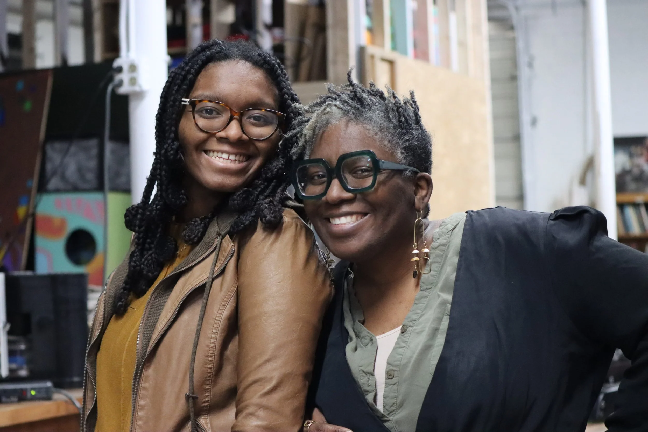 Two smiling women with glasses, one with braided hair and the other with dreadlocks, posing together in a room filled with shelves and books.