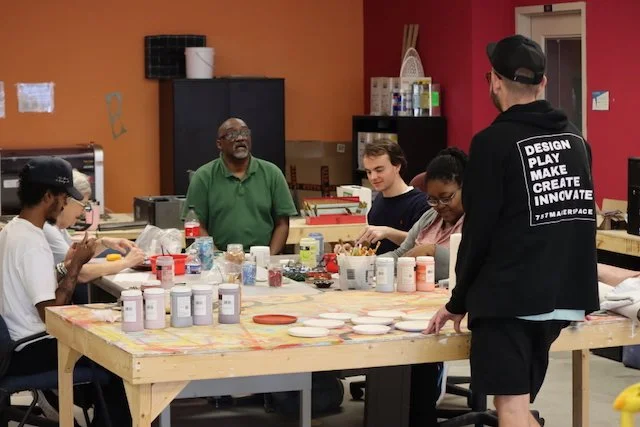 Group of people in a ceramics or pottery class, sitting and standing around a large work table with paint and pottery supplies, in a colorful art studio.